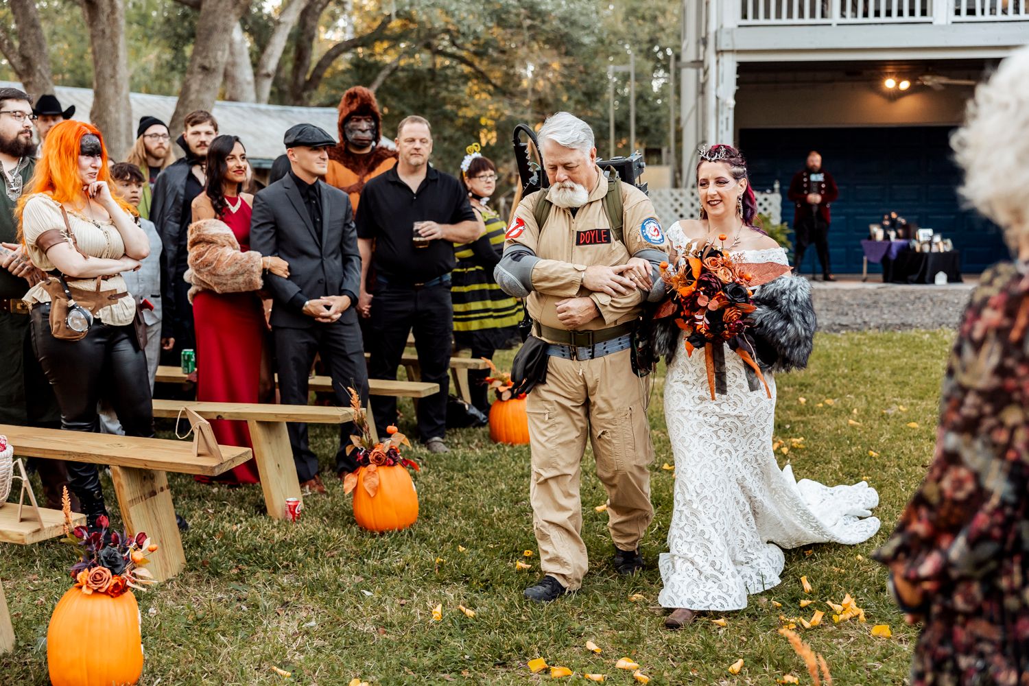 Guests gather outdoors around wooden benches decorated with autumn pumpkins during a fall wedding ceremony.