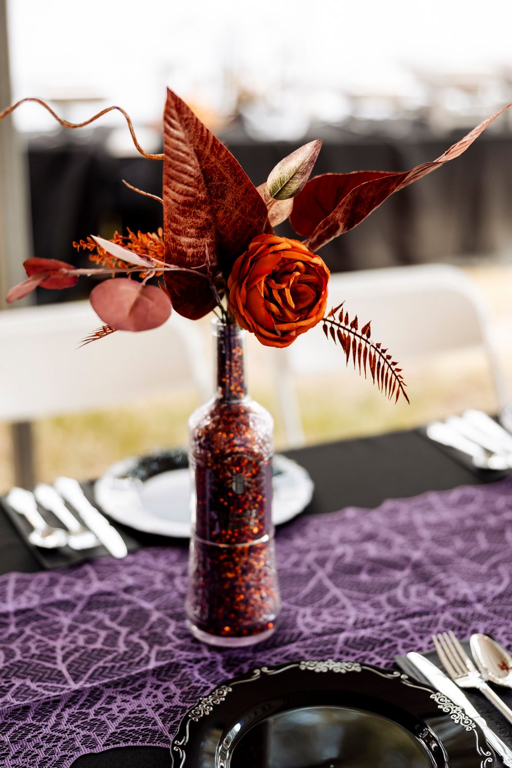 Red and copper autumn floral arrangement in glitter-covered bottle on purple lace tablecloth.