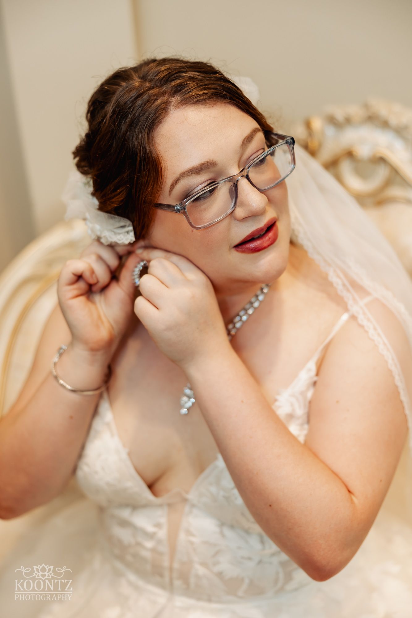 A bride in glasses and red lipstick adjusts her earrings while wearing an elegant lace wedding gown.
