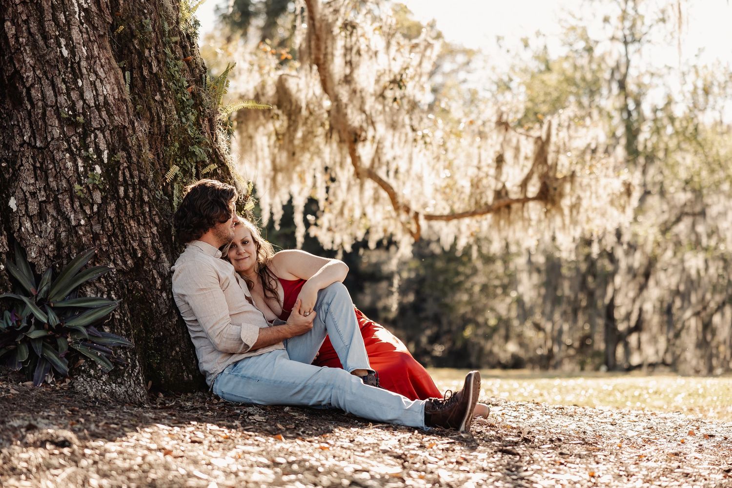 A romantic couple sits together leaning against a tree, reading a book in a sunlit park.