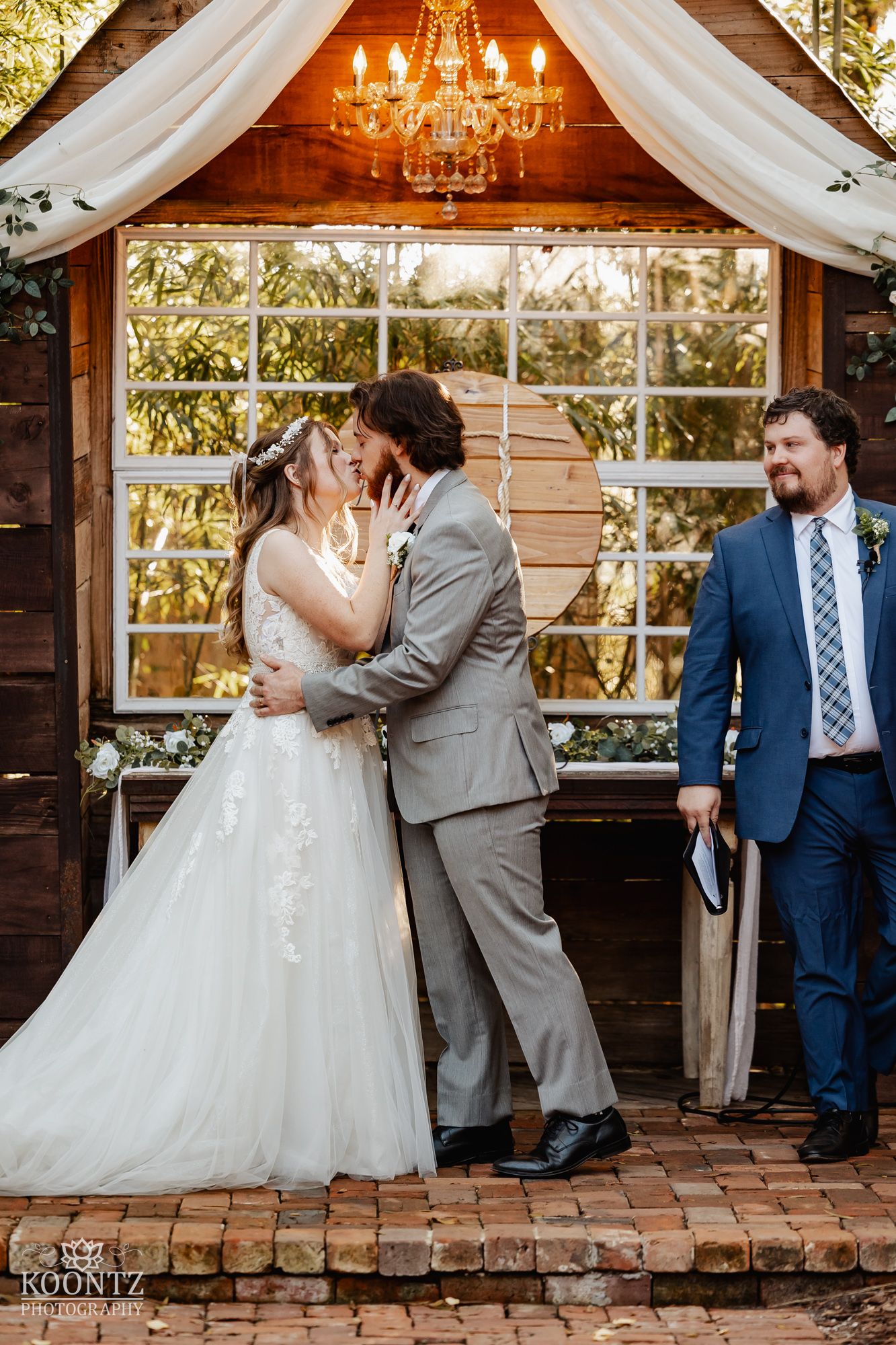 Bride and groom share first kiss at rustic chandelier-lit wooden altar as officiant smiles nearby.