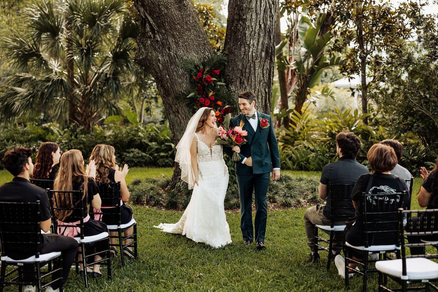 Newlyweds face guests after outdoor wedding ceremony surrounded by lush greenery and red floral decor.