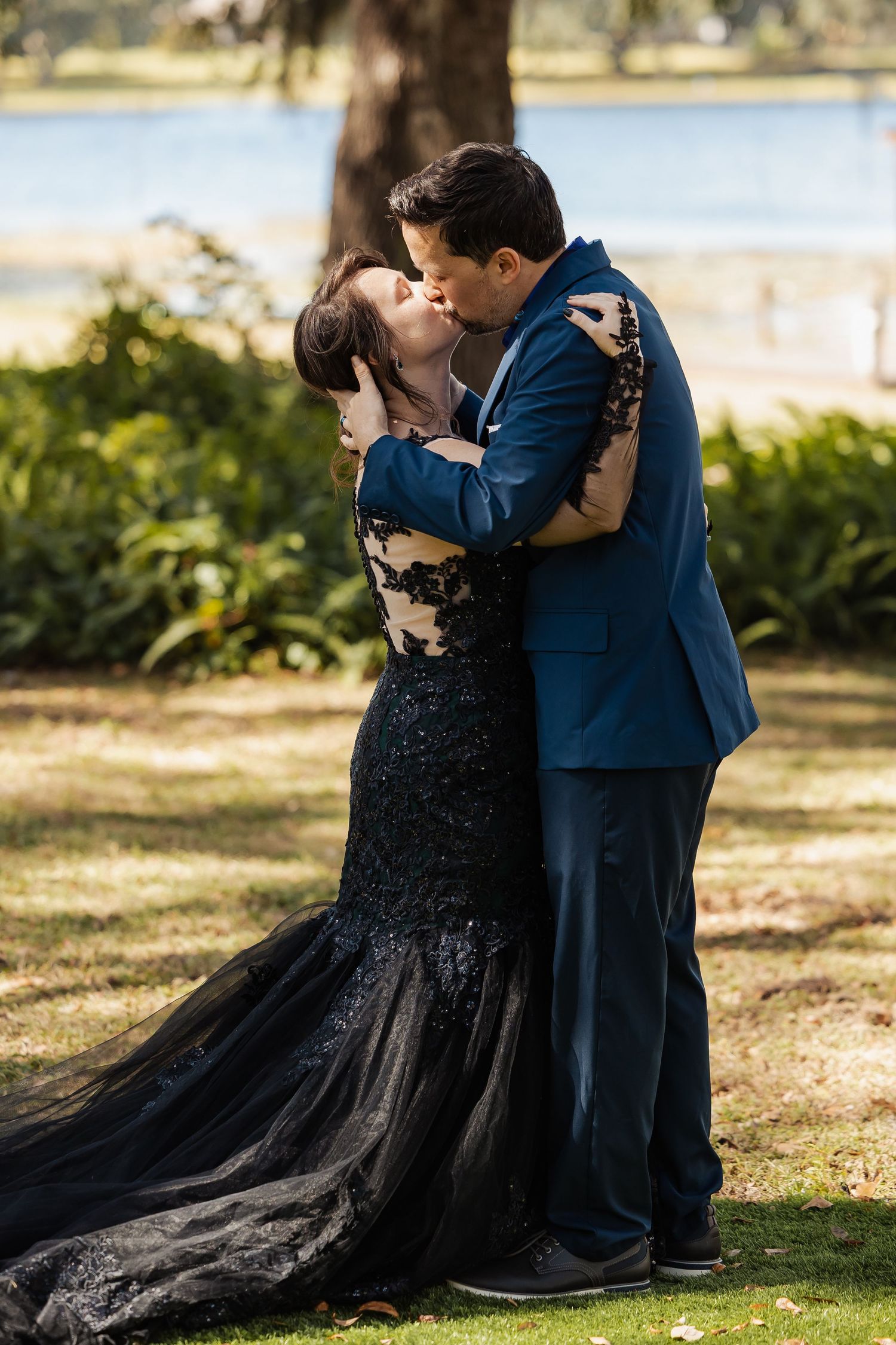 Newlyweds share first kiss in black lace gown and navy suit beside a serene lake at outdoor wedding.