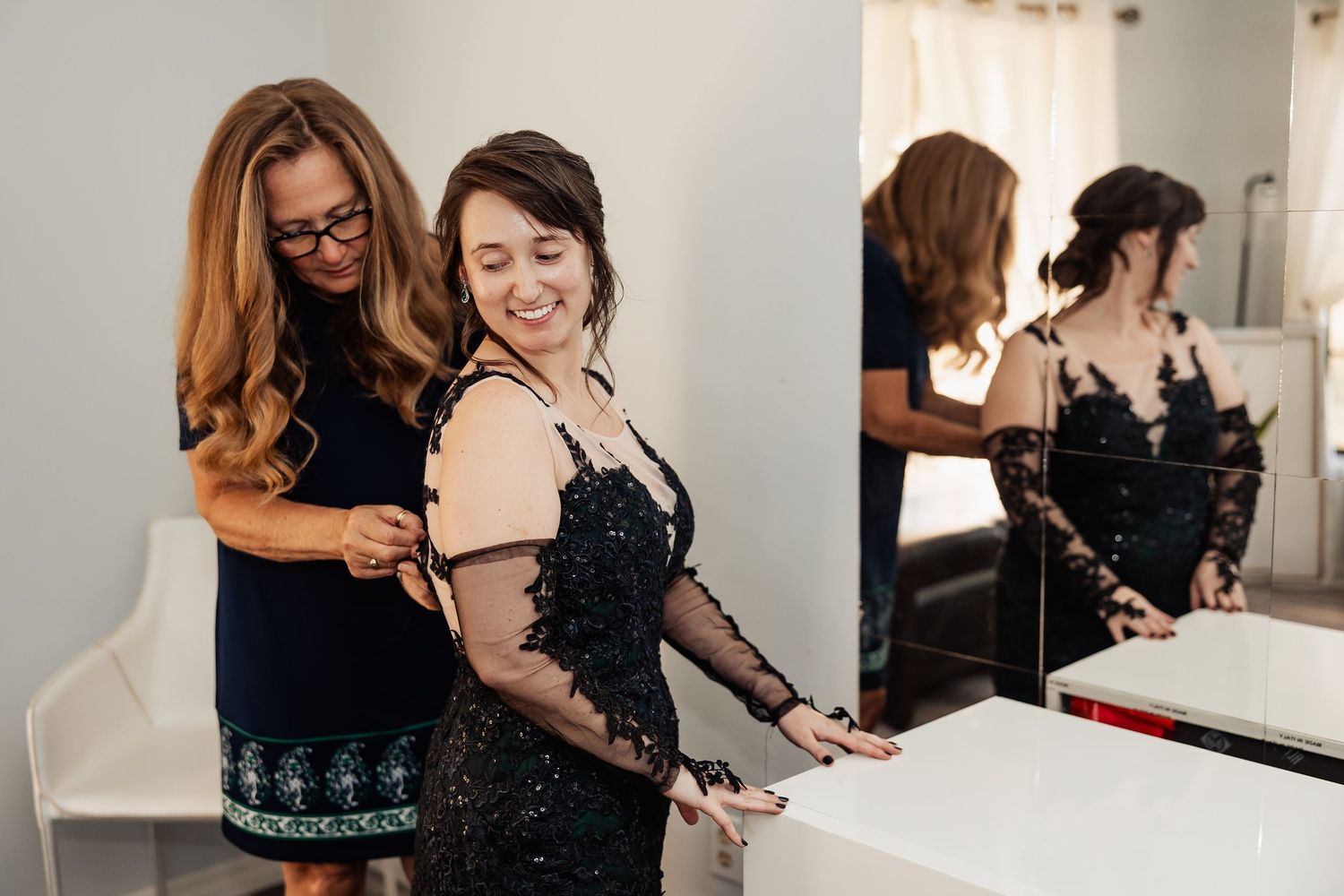 Woman in black lace dress smiling as bridesmaid adjusts her gown near mirror before wedding.