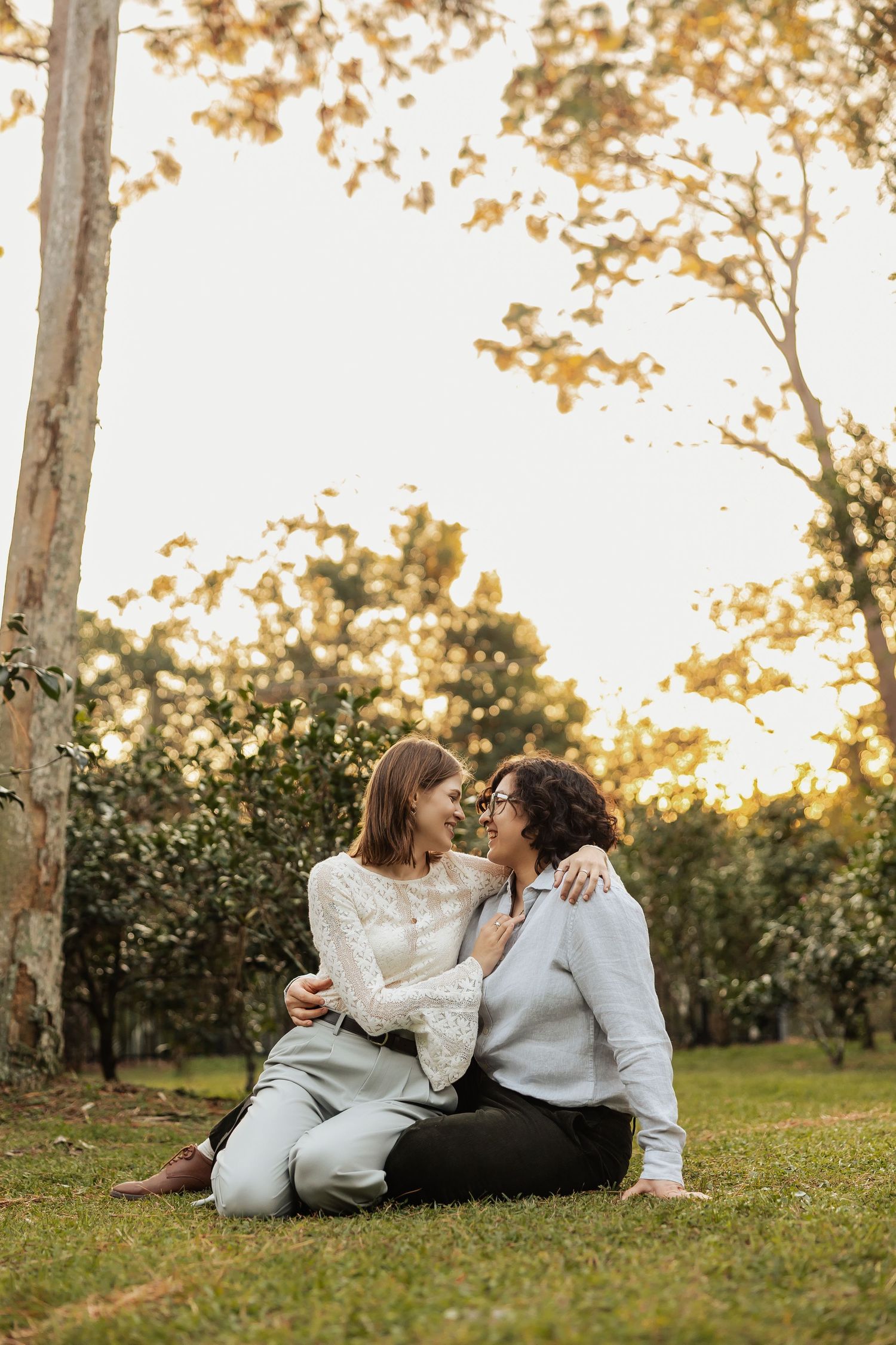 Two women sitting on grass in a sunlit park, smiling and embracing each other warmly.