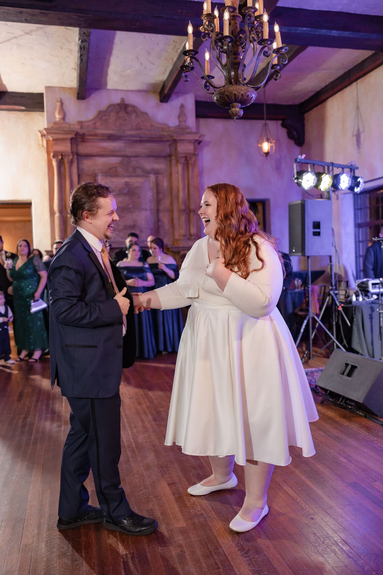 Bride in white dress laughing with groom during first dance at elegant wedding reception with chandelier.