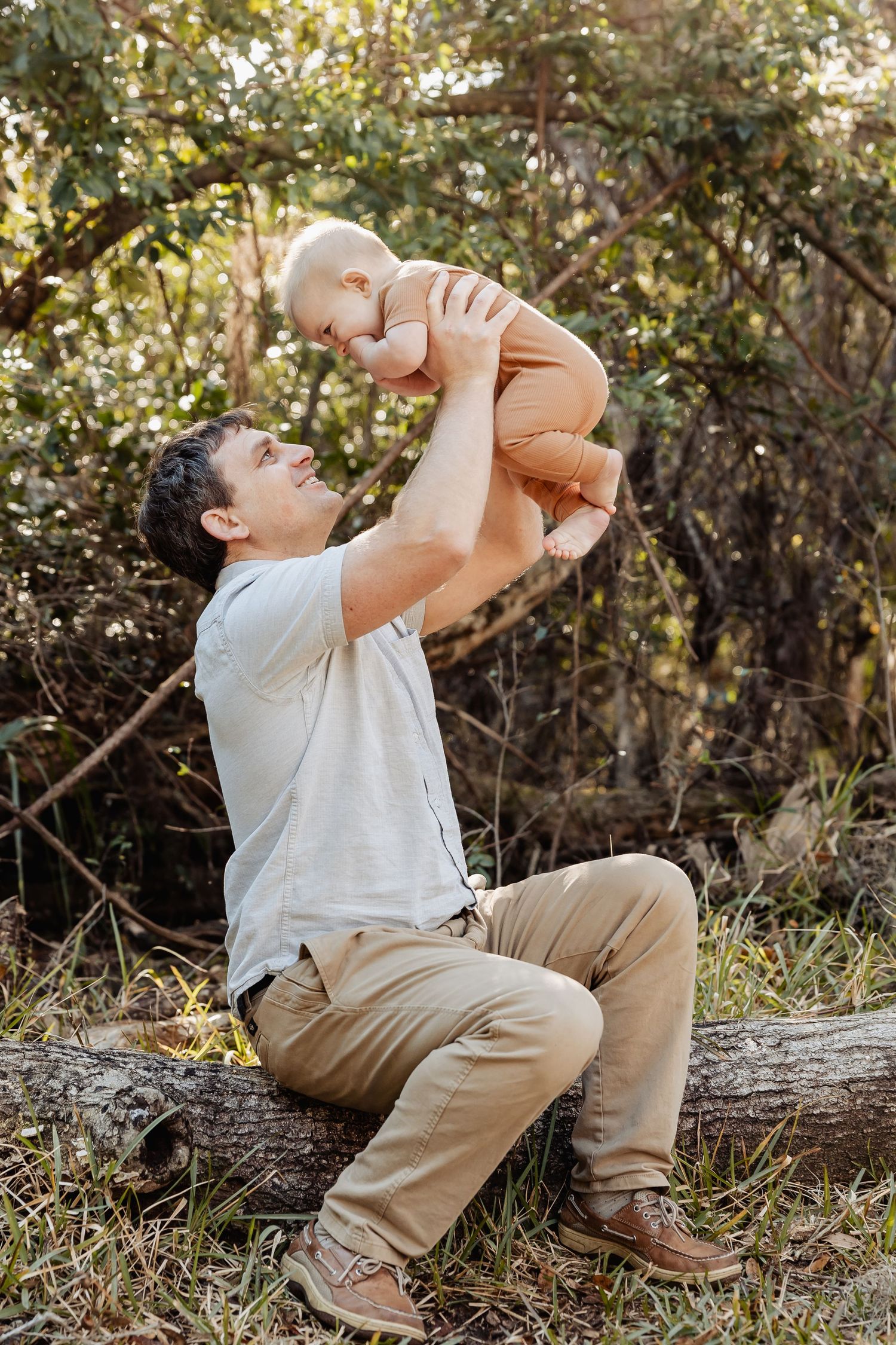Father sitting on a log lifting his smiling baby in the air outdoors in a natural setting.