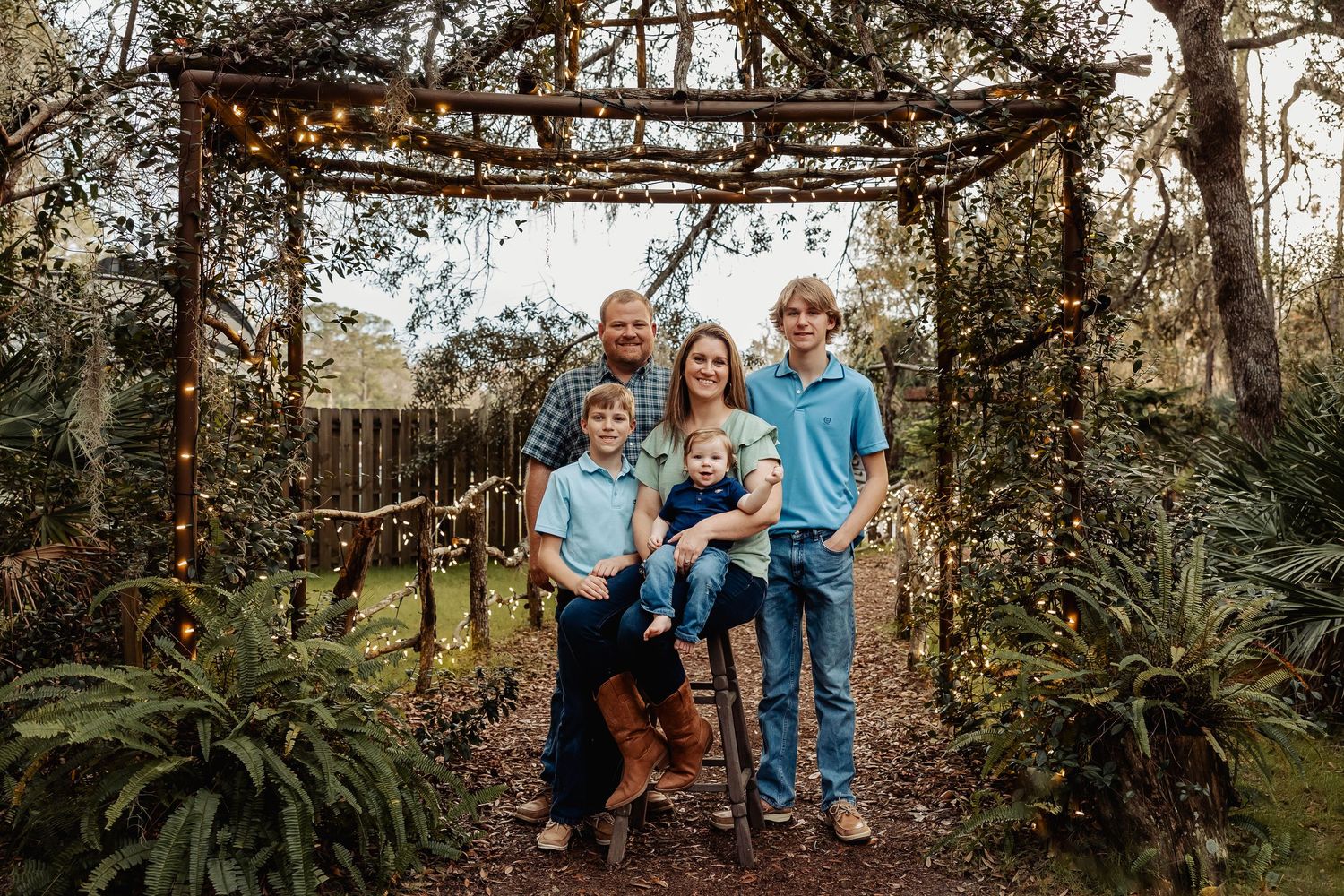 Happy family of five posing under a rustic garden arbor with string lights surrounded by greenery.