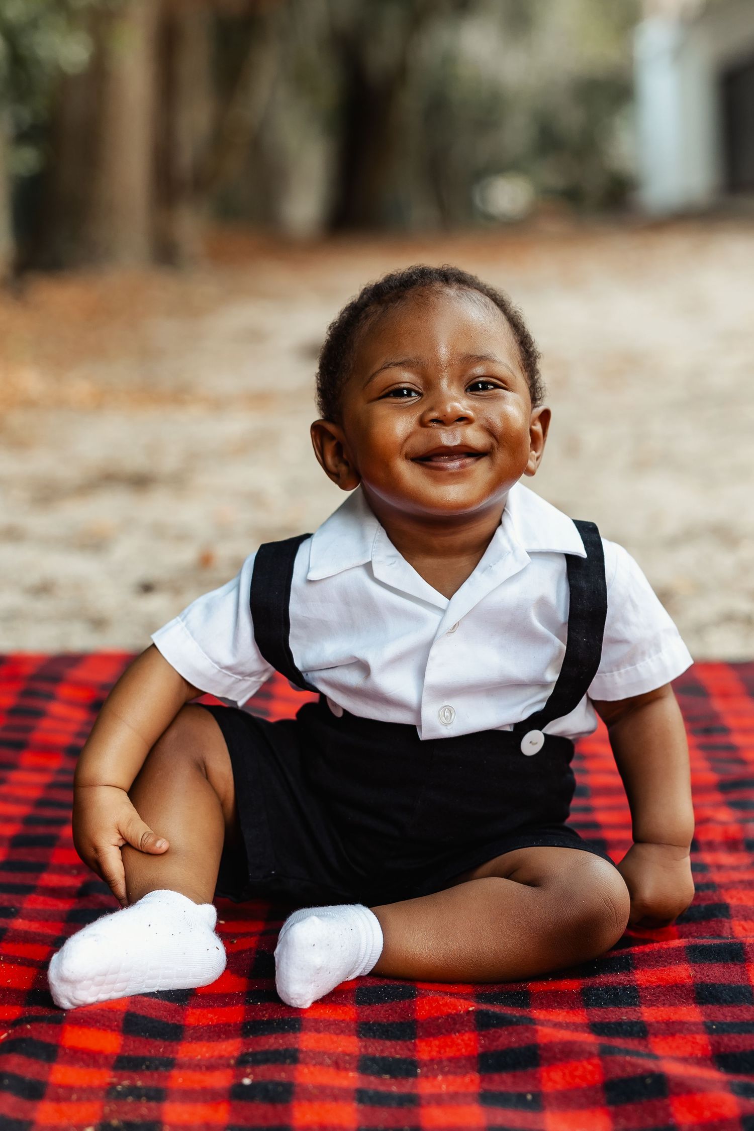 Adorable smiling Black baby boy in suspenders sitting on red plaid blanket outdoors.
