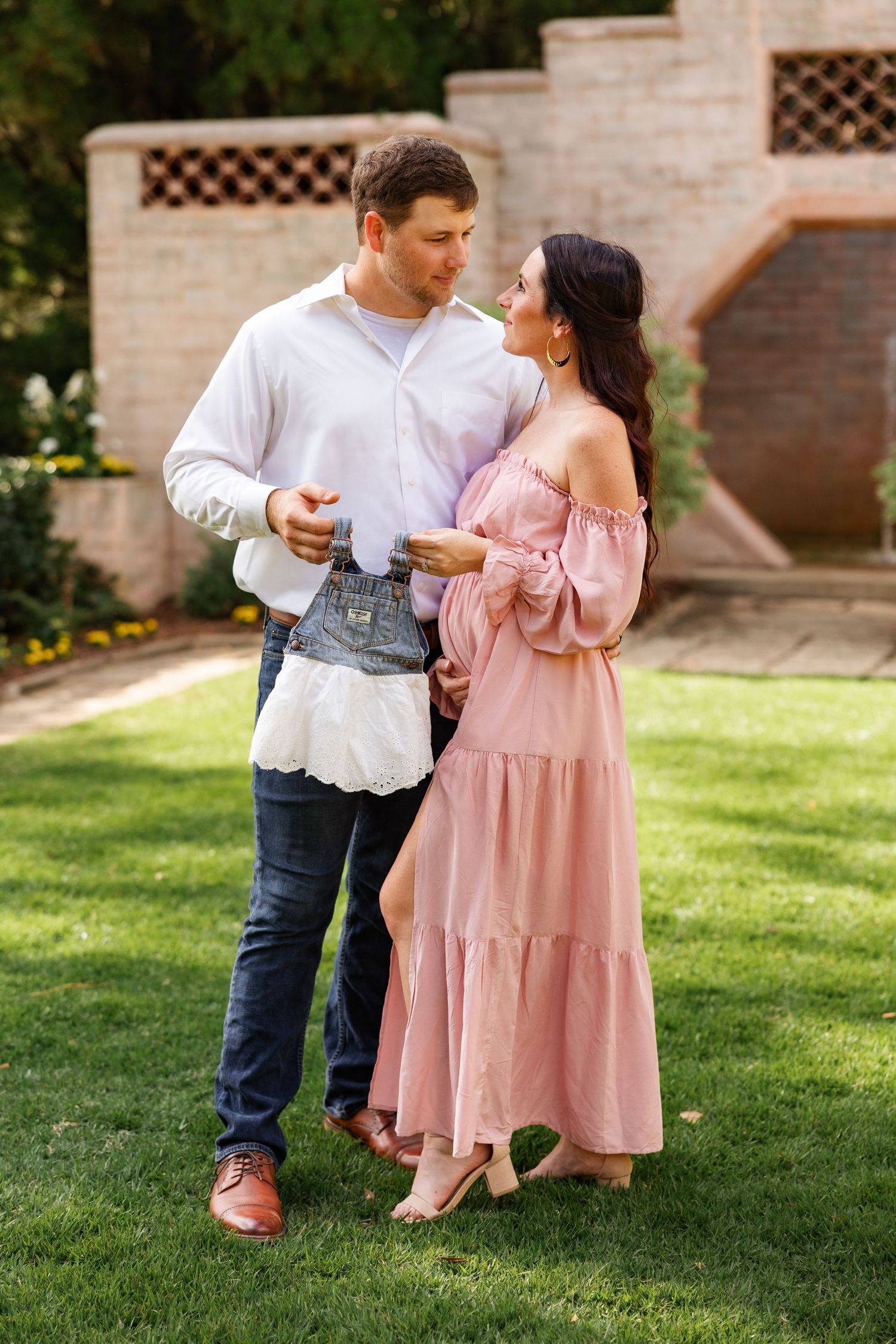 Expectant couple standing on green lawn holding tiny baby clothes, woman in pink off-shoulder dress, man in white shirt.