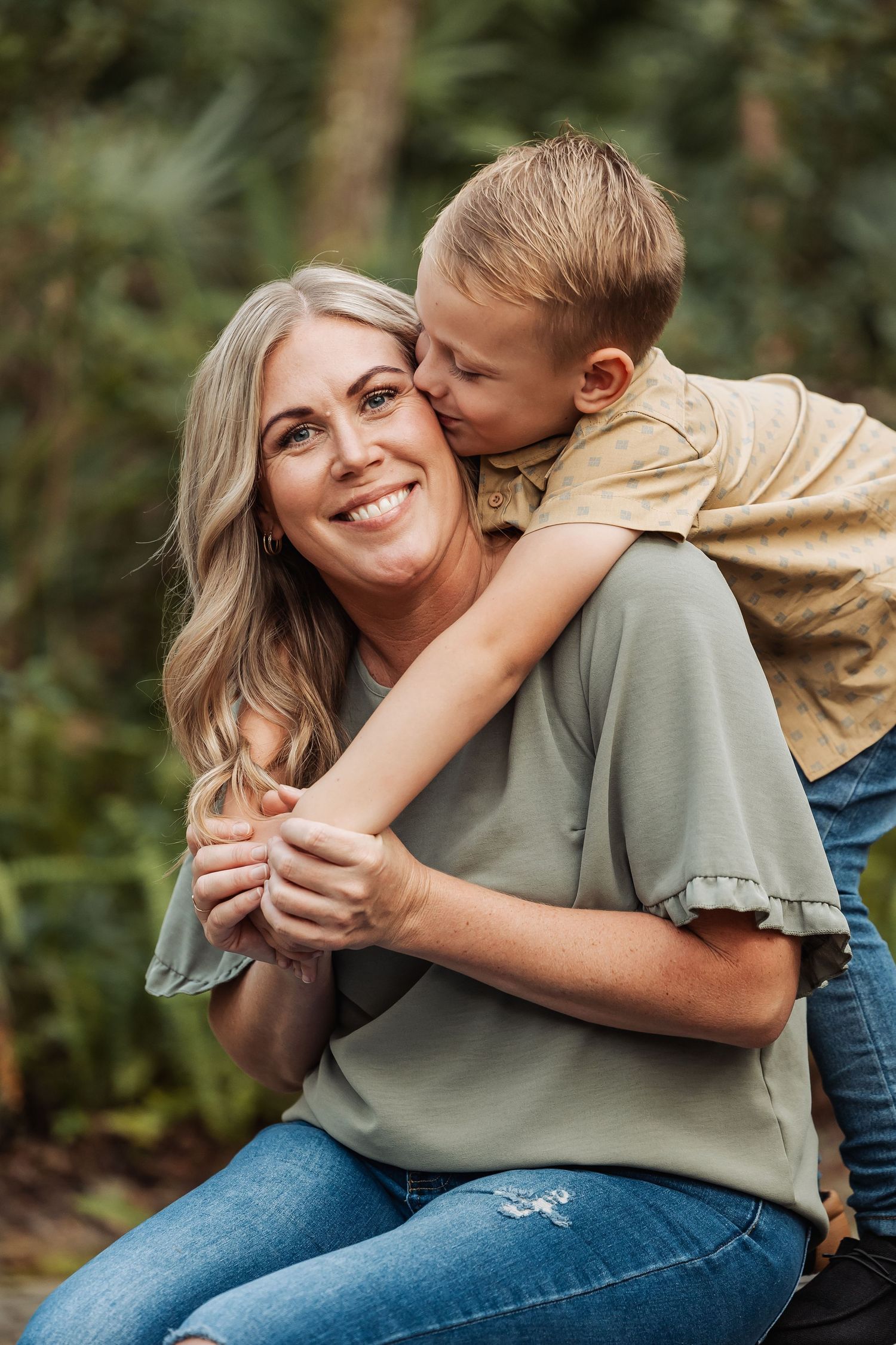 A young boy hugs and kisses his smiling mother on the cheek in a green outdoor setting.