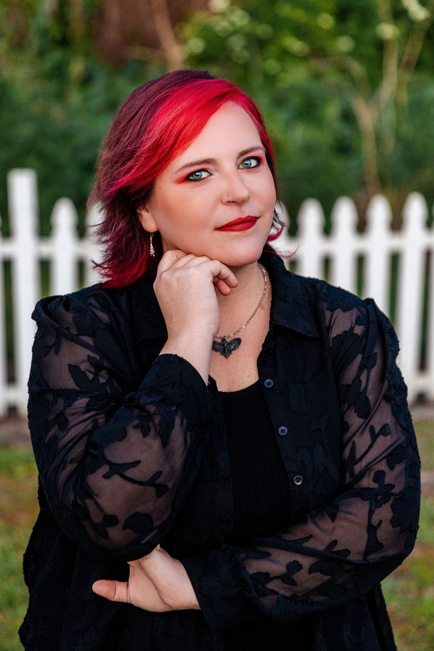 Woman with vibrant red hair in black lace blouse poses thoughtfully against white picket fence outdoors.