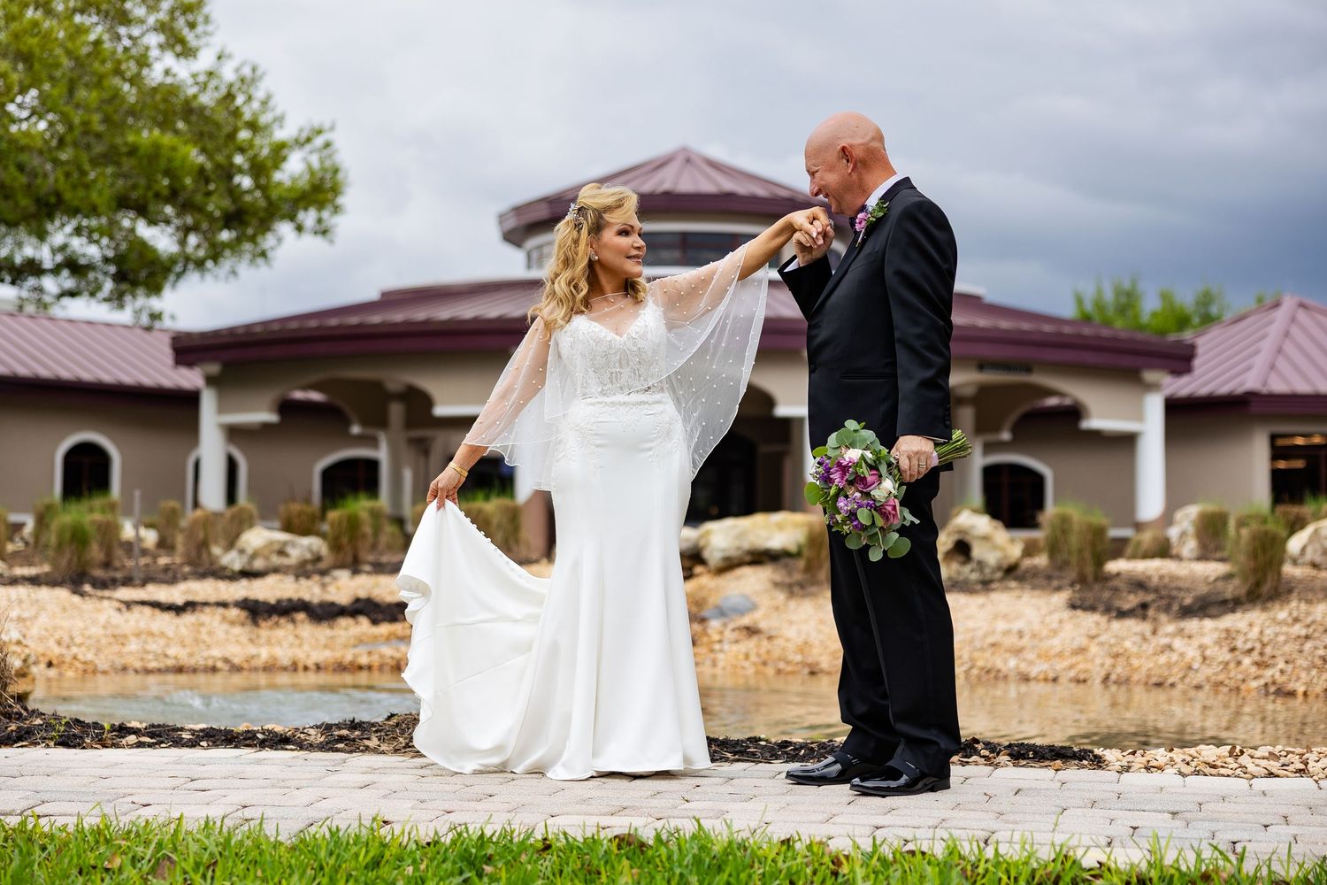 Groom kissing bride's hand in white gown outdoors near pond at elegant wedding venue with lush greenery.