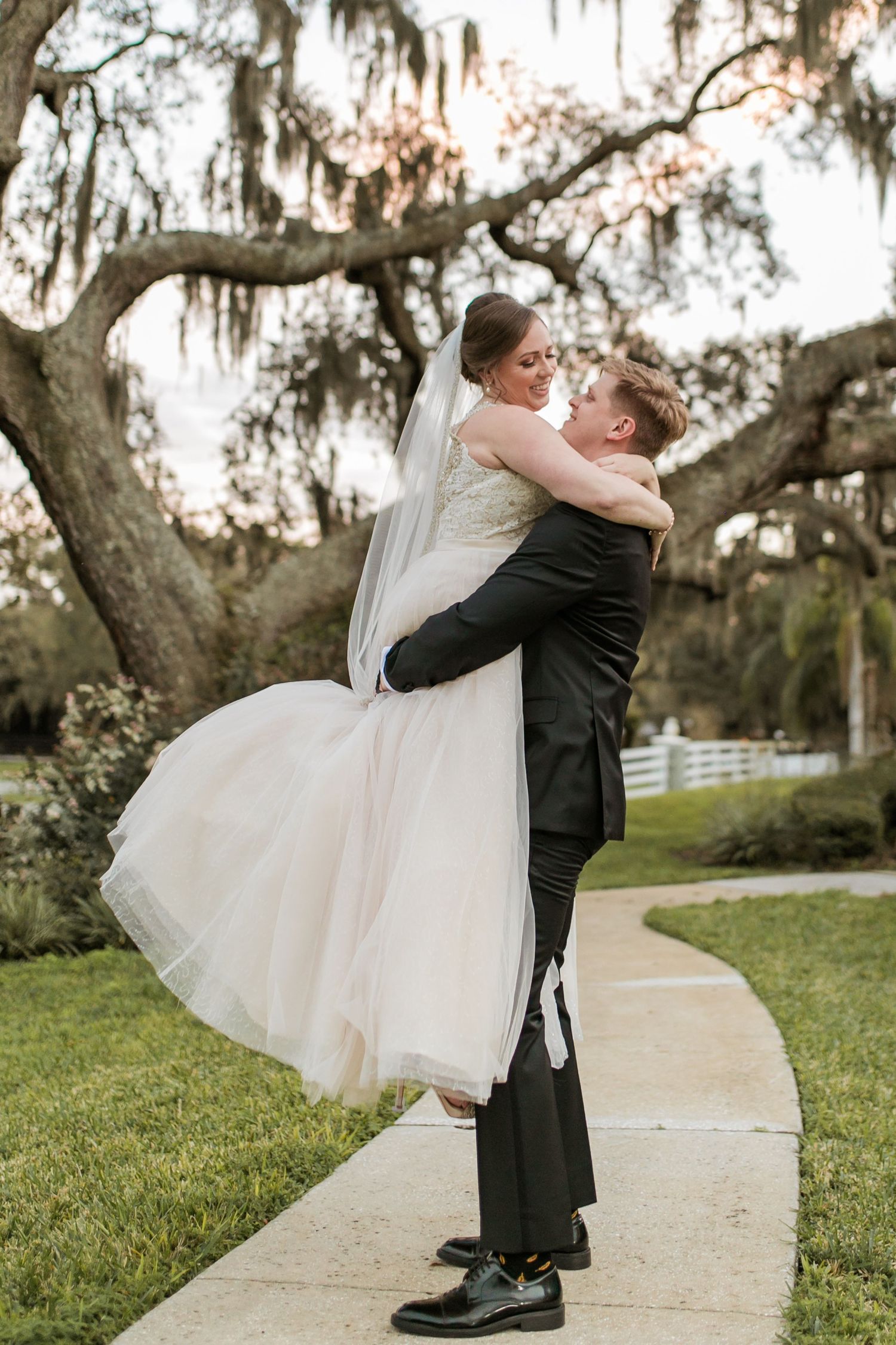 Groom lifts bride in white gown on garden path, sharing a romantic moment beneath a sprawling moss-covered oak tree.