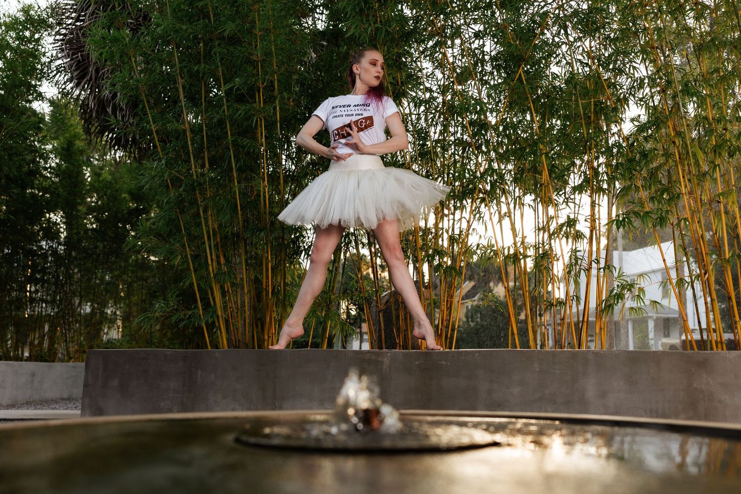 Ballerina in white tutu and graphic tee standing gracefully near a fountain with bamboo background.