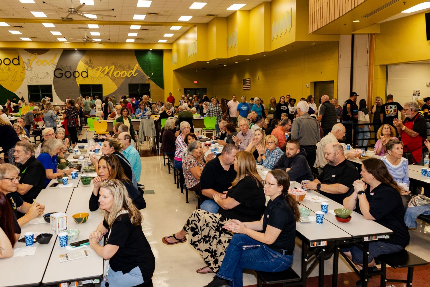 A large crowd of attendees sit at tables in a decorated cafeteria during a community gathering.