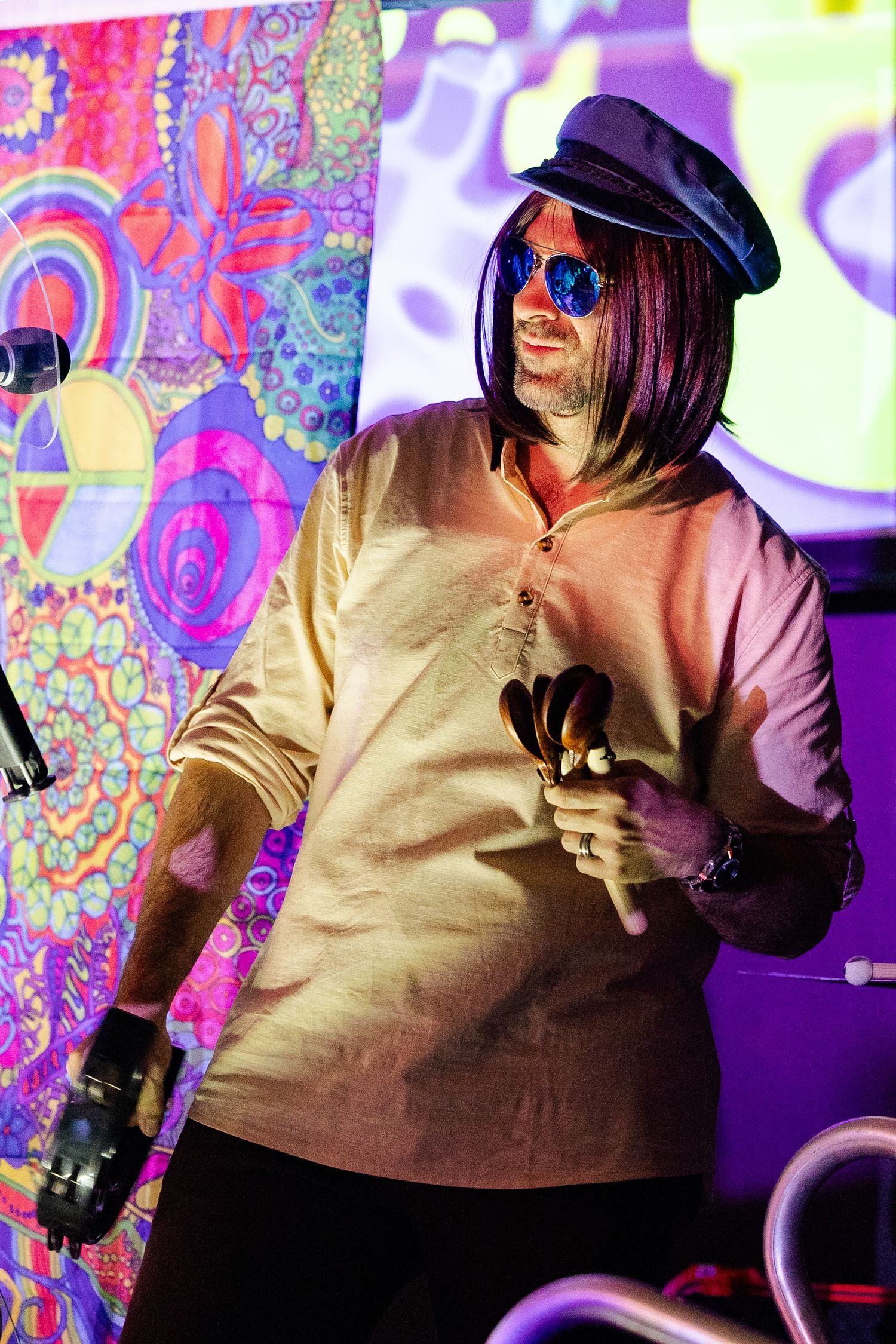 Male performer in 1960s-style outfit with sunglasses and cap playing castanets on a colorful stage.