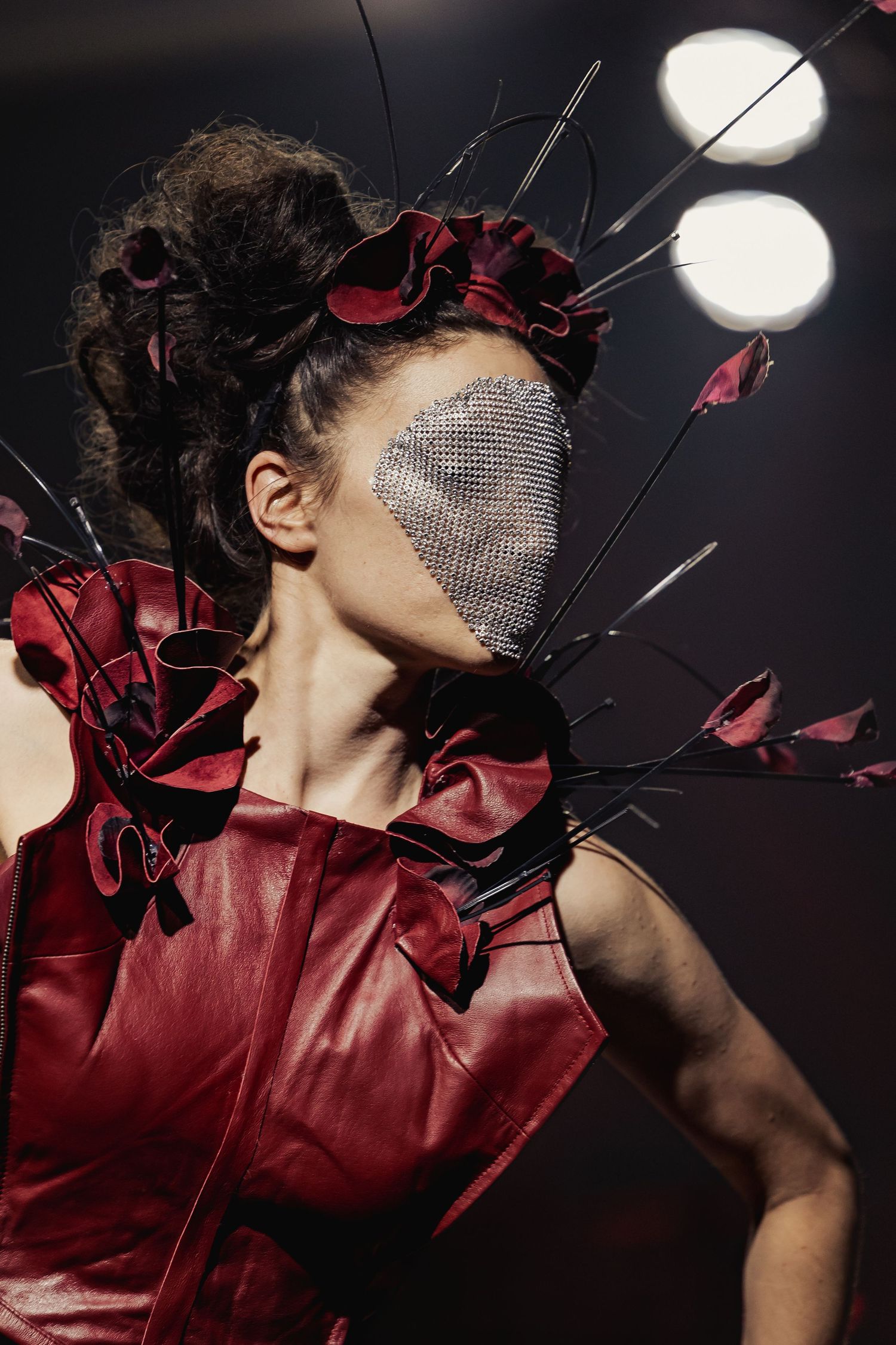 Close-up of model in red leather top with floral headpiece and silver mesh face mask at fashion show.