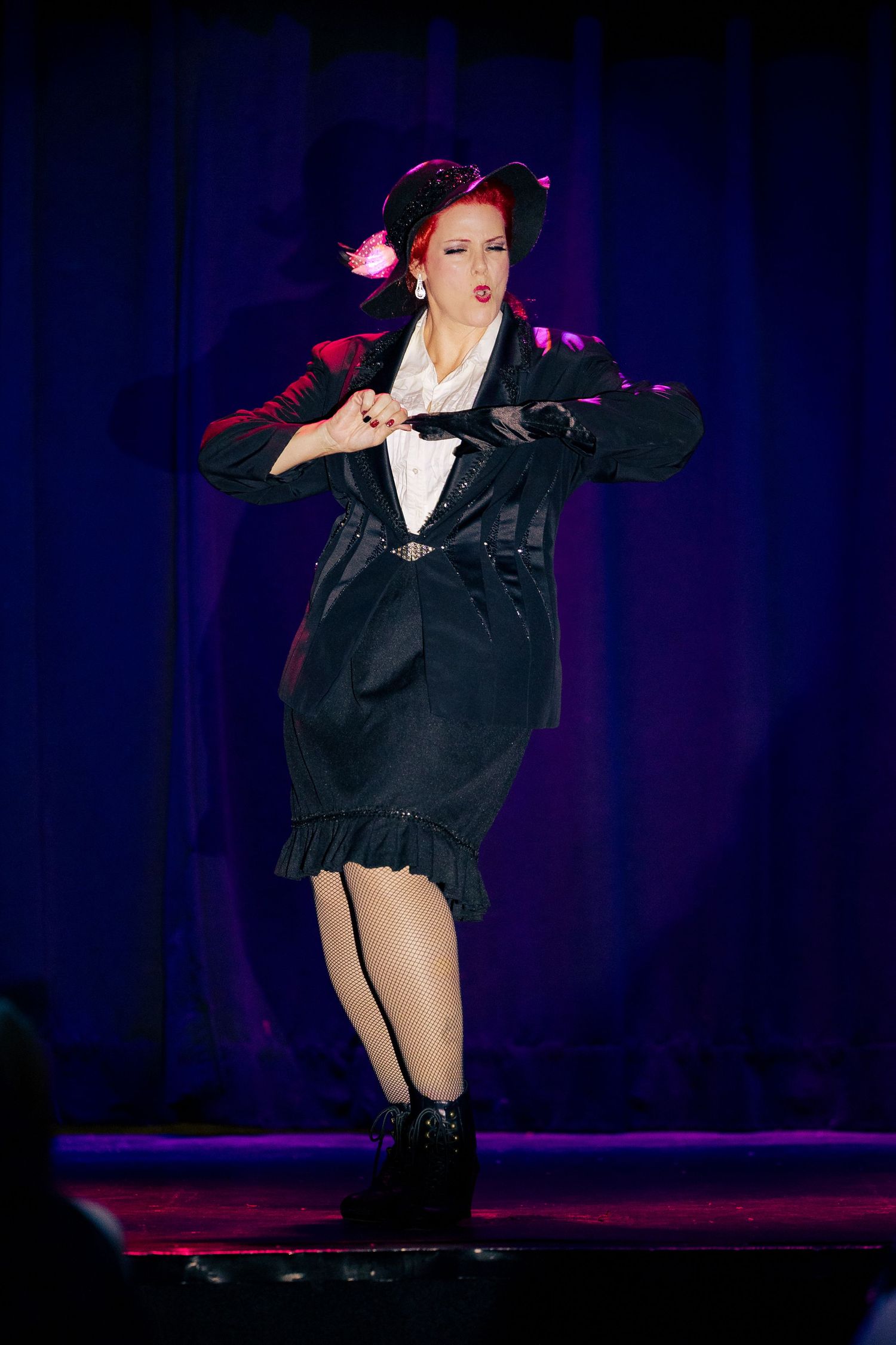 A redhead performer in a black dress adjusts her costume on a colorfully lit burlesque stage.