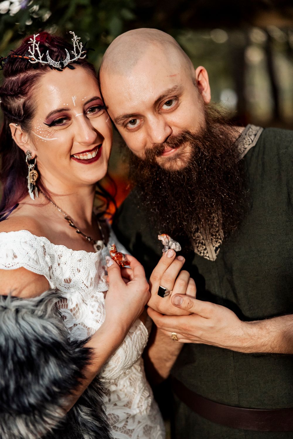 Viking bride with antler crown and fantasy makeup poses with bearded groom holding small animal figurines.