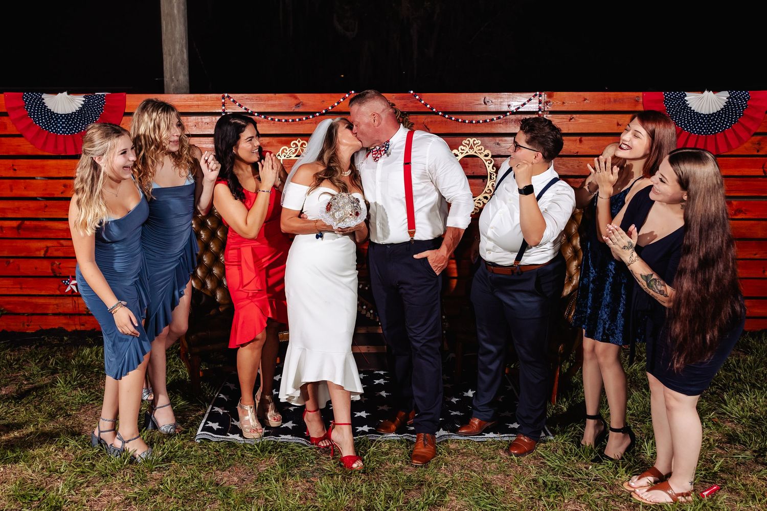 Bride and groom pose with wedding party guests in front of patriotic decorated wooden wall at reception.