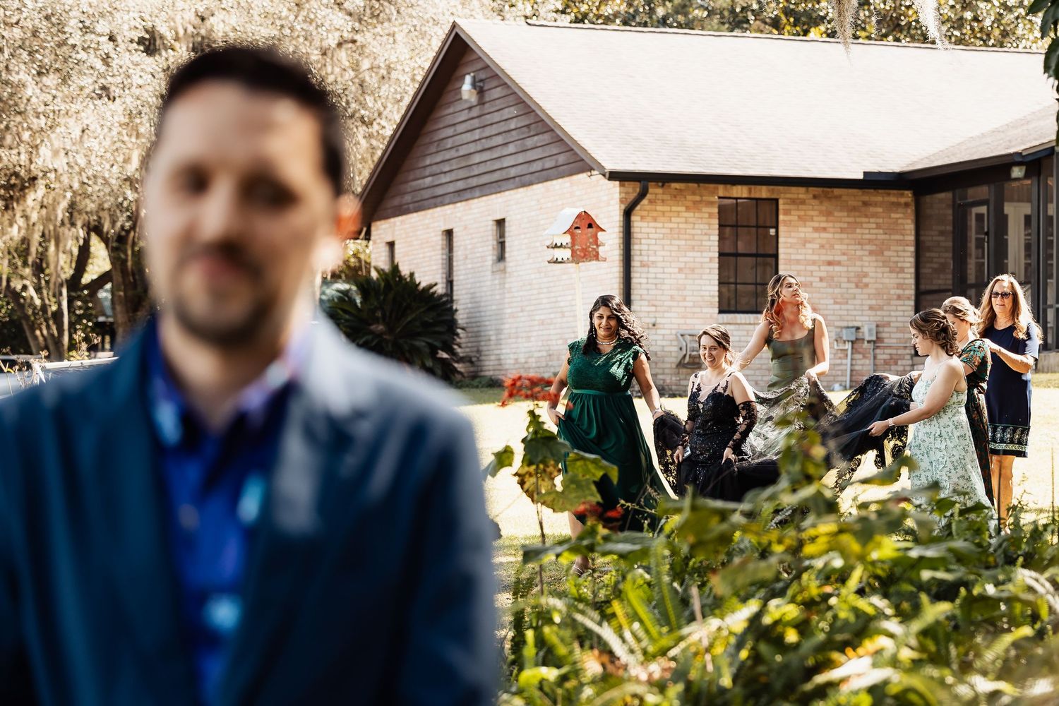 Groom in blue suit stands in foreground as wedding party gathers outside rustic brick venue.