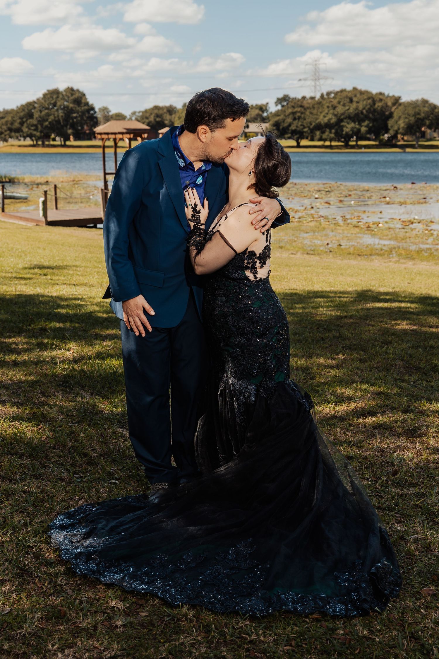 Newlyweds kiss passionately in black lace gown and navy suit beside a peaceful lake after outdoor ceremony.