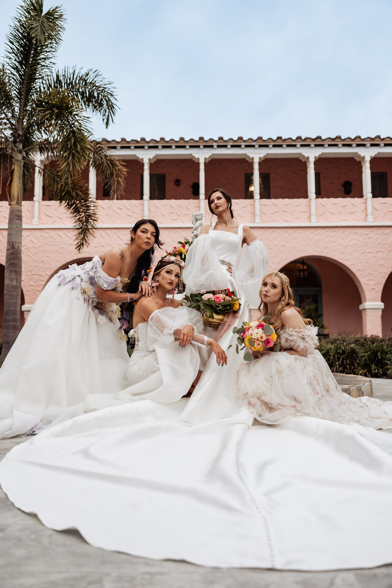 Four brides in elegant white wedding gowns pose together outside a pink Spanish-style venue.