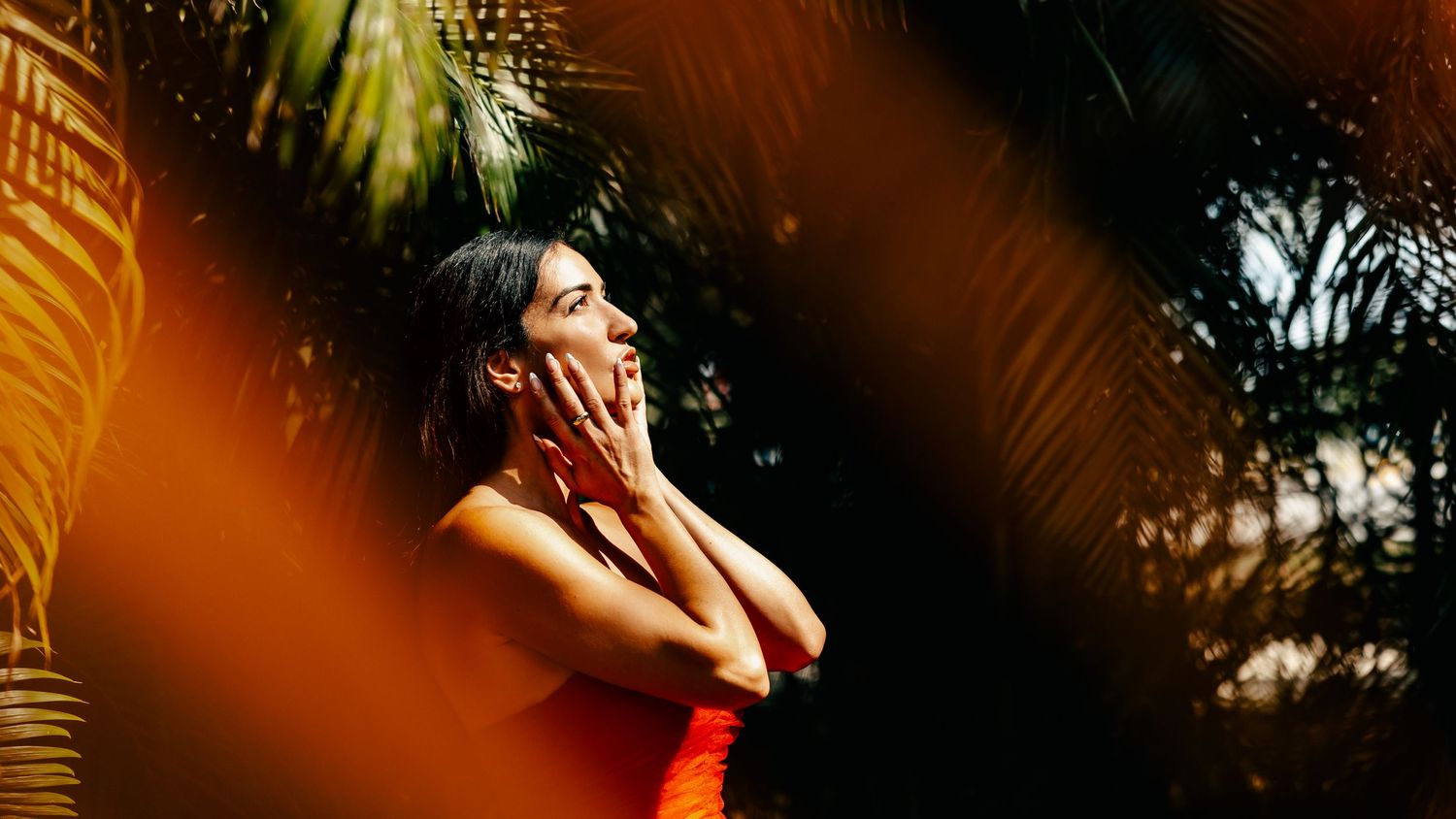 Artistic portrait through palm leaves with warm orange light and shadows.