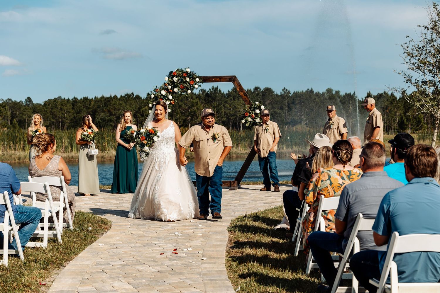 An outdoor wedding ceremony takes place on a scenic lakeside path with white chairs and decorative wooden arch.