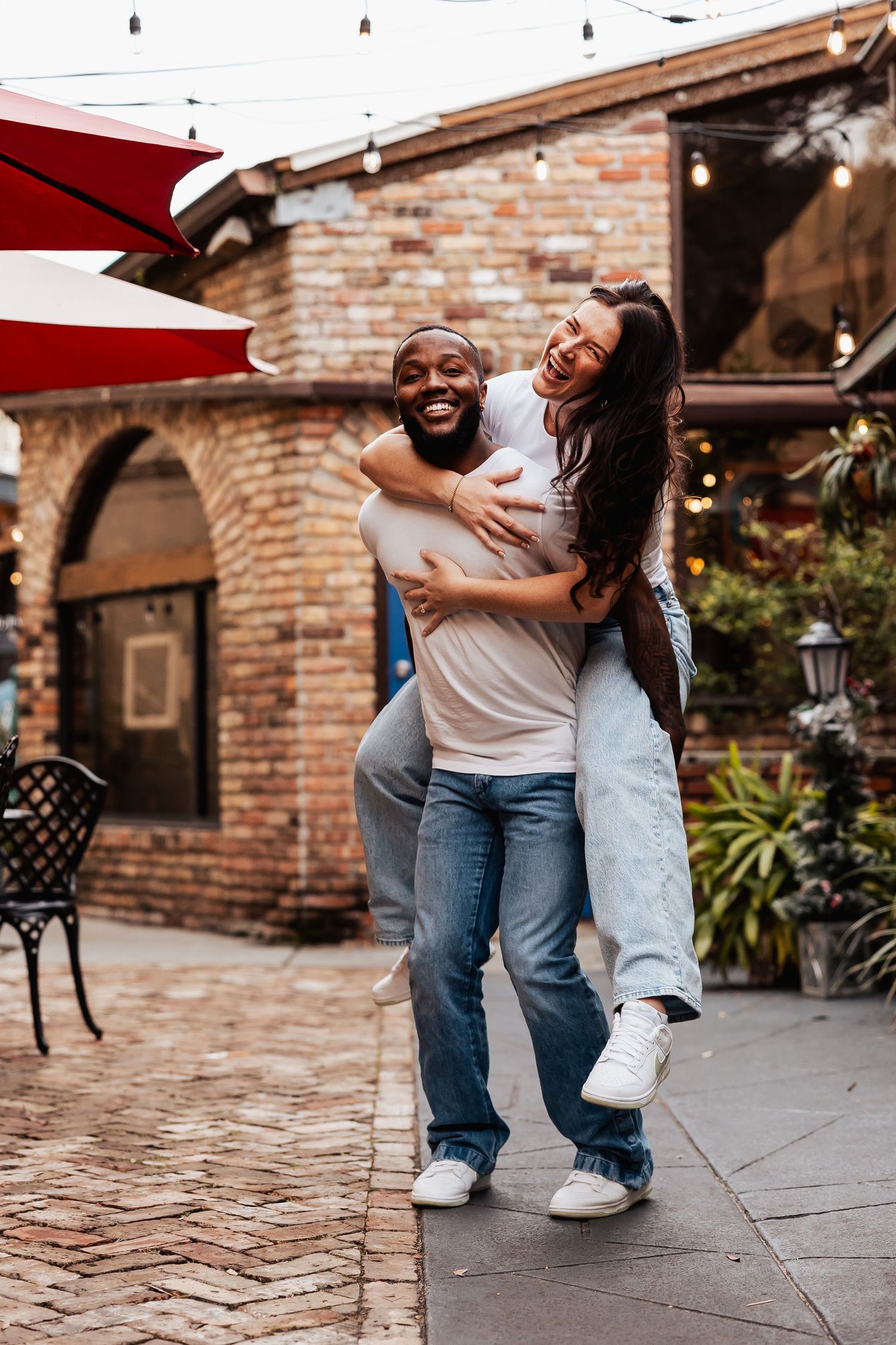 Playful piggyback moment shared between a couple in a charming brick courtyard with red umbrellas.