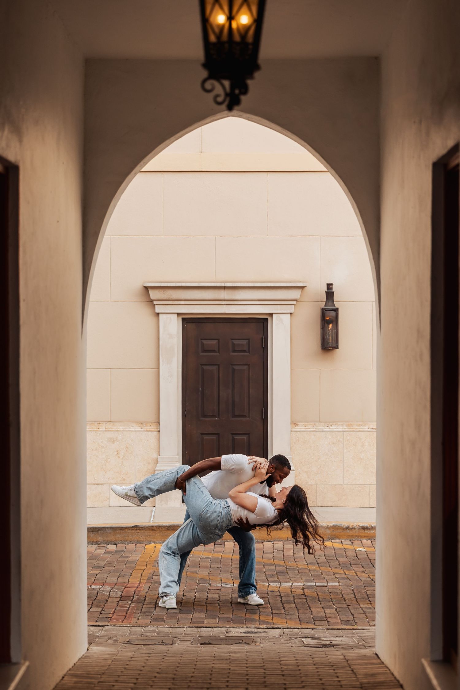 Romantic couple performs playful dance dip pose under architectural archway with vintage lantern.