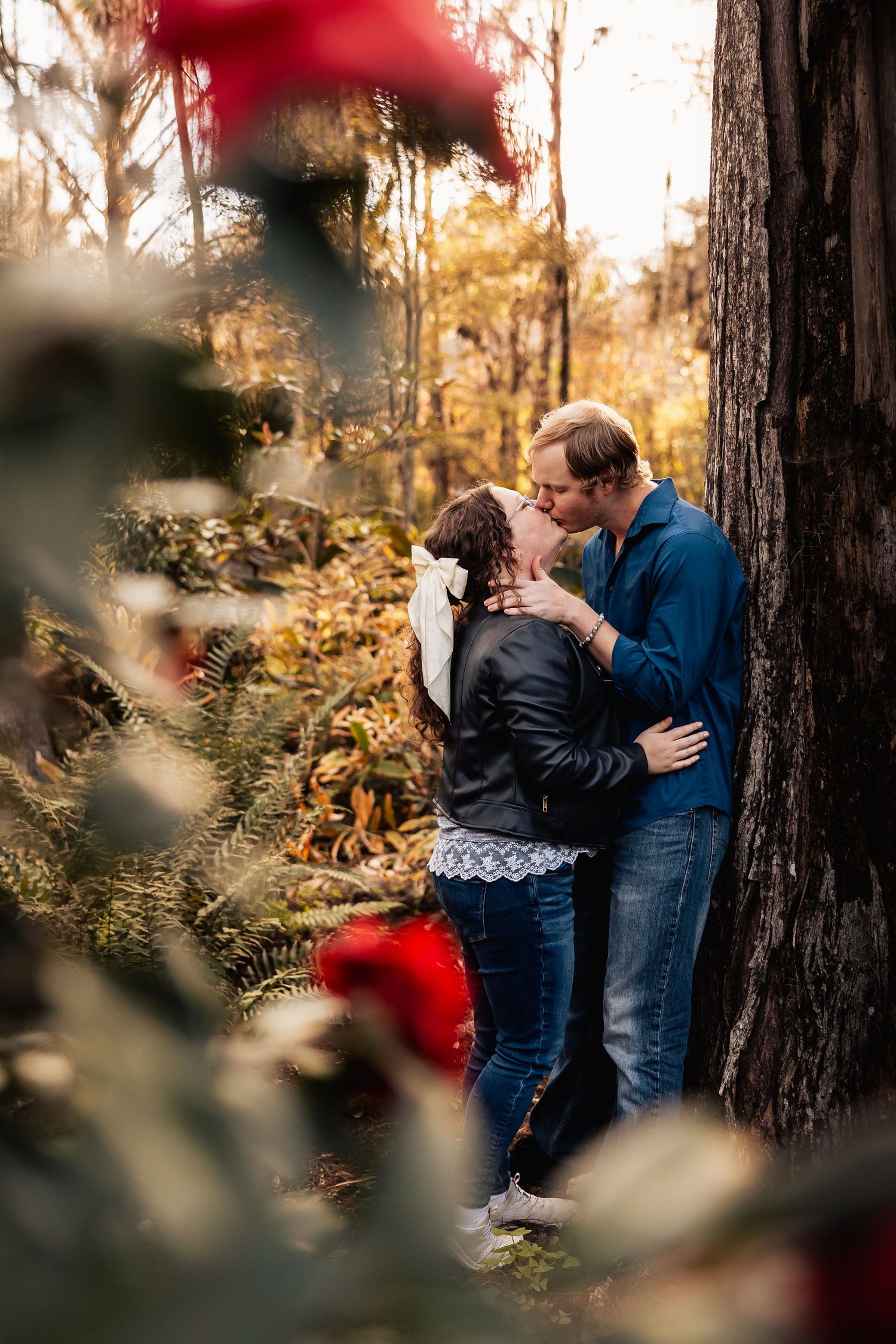 Couple sharing a romantic kiss against a tree surrounded by autumn ferns and foliage.