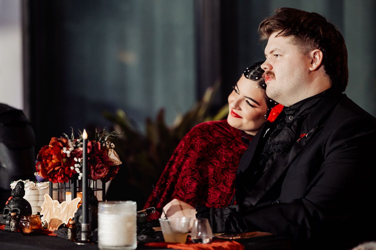 Couple in gothic wedding attire sitting together at a decorated table with red flowers and candles.