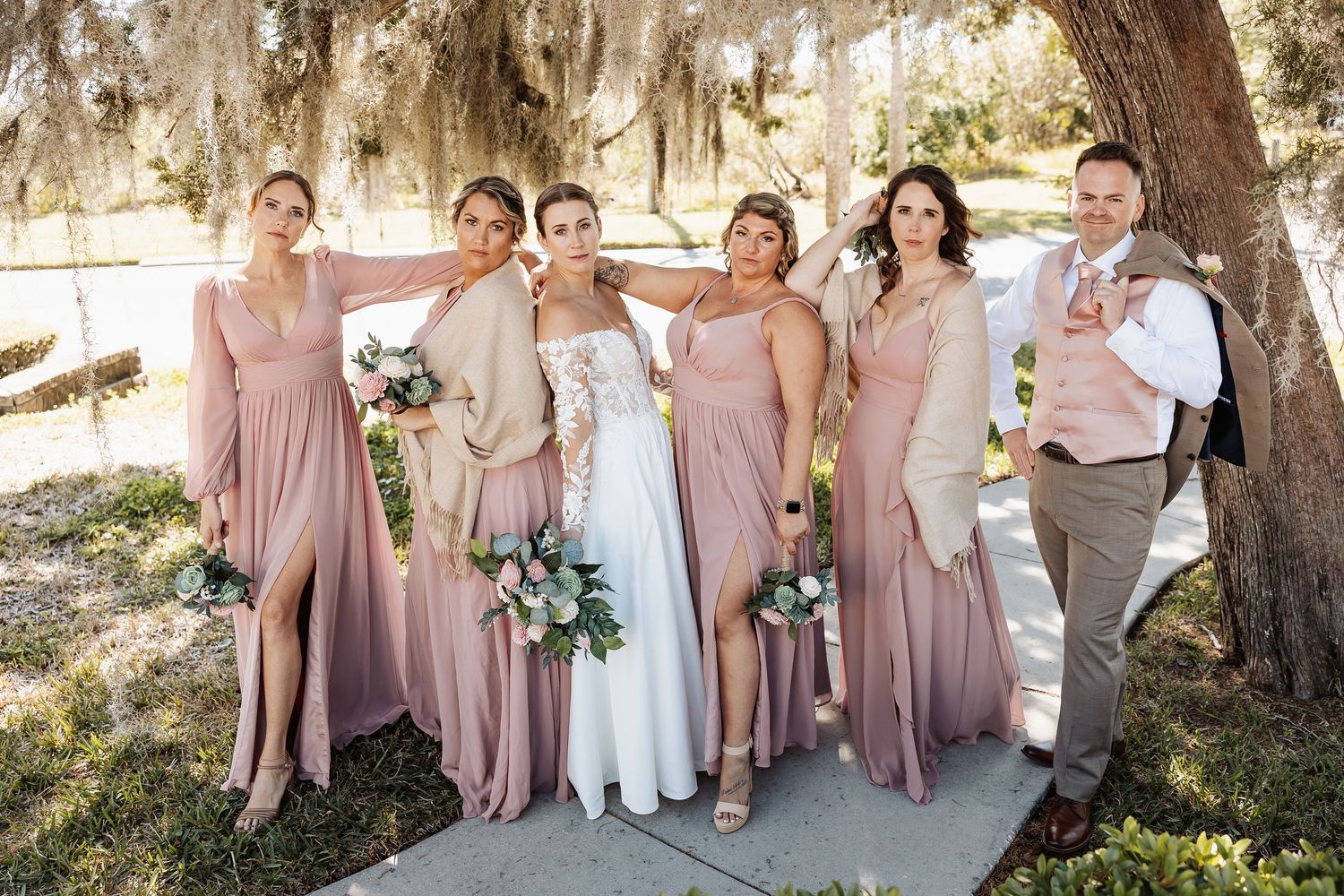 Bride poses with four bridesmaids in dusty rose gowns and a groomsman outdoors under a large oak tree.