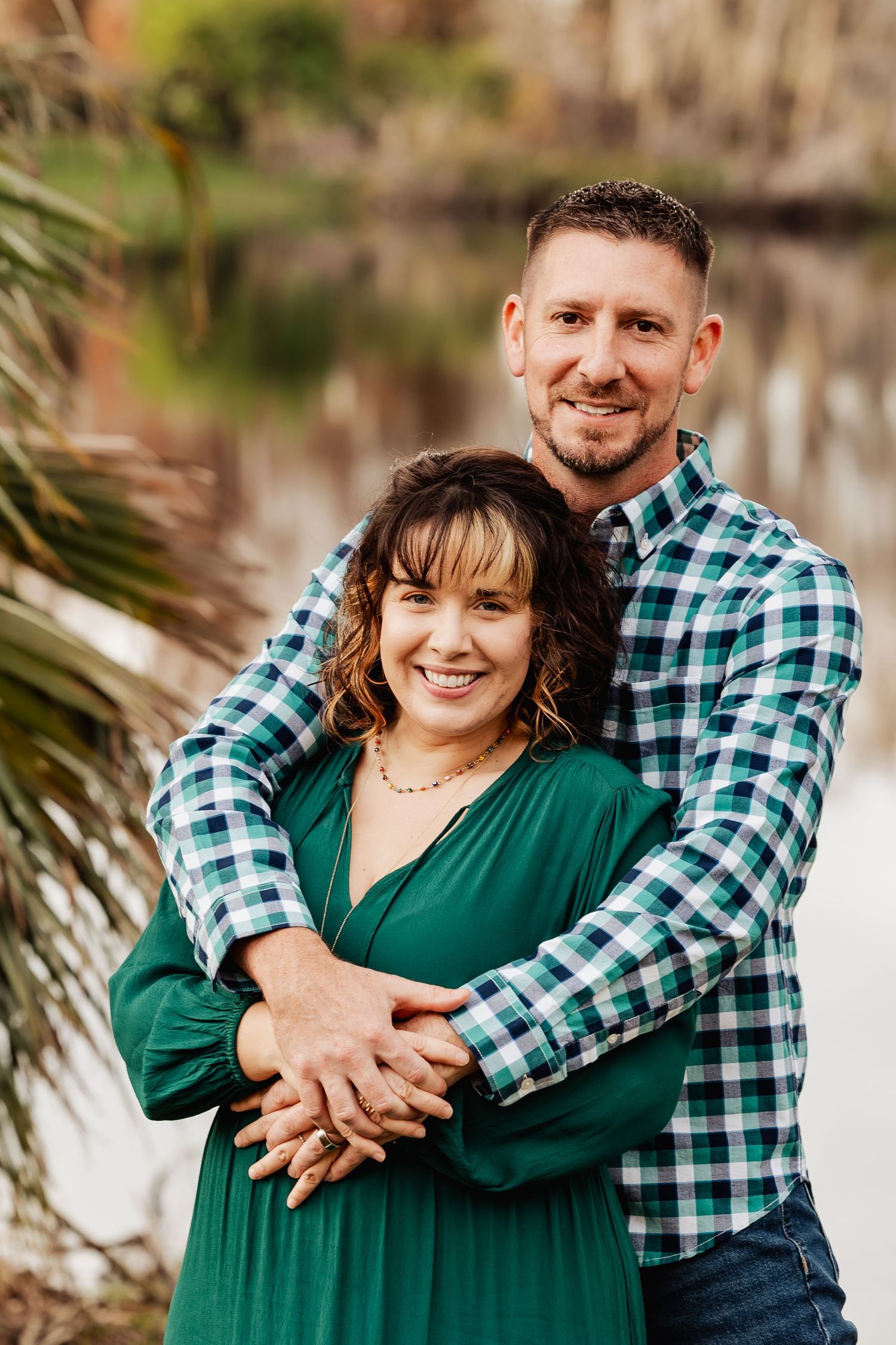 Happy couple embracing outdoors, woman in green dress and man in plaid shirt smiling by lake.