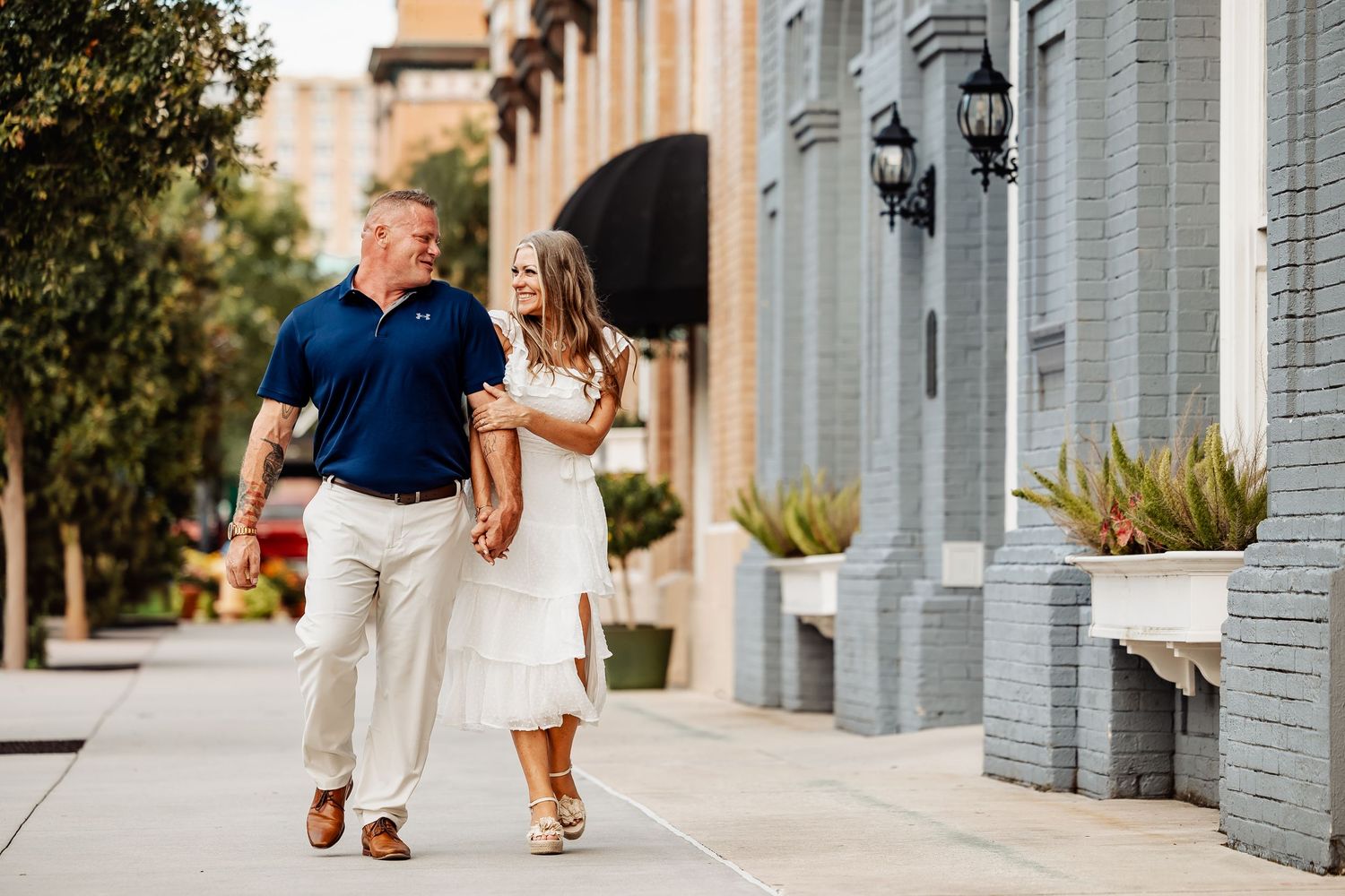 Happy couple walking hand in hand along a sunny urban sidewalk, woman in white dress, man in navy polo.