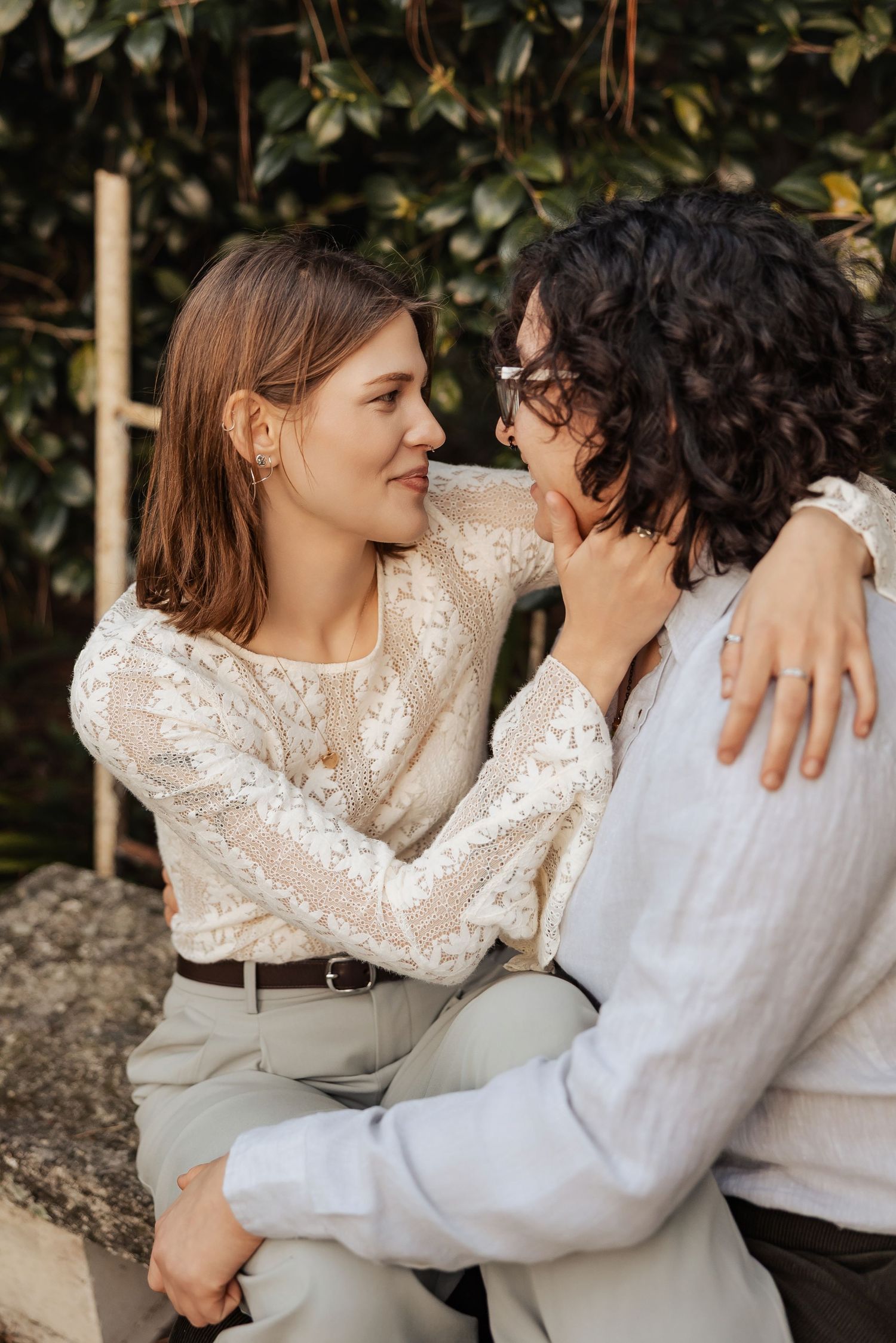 Two women sitting closely outdoors, one tenderly touching the other's face while smiling.