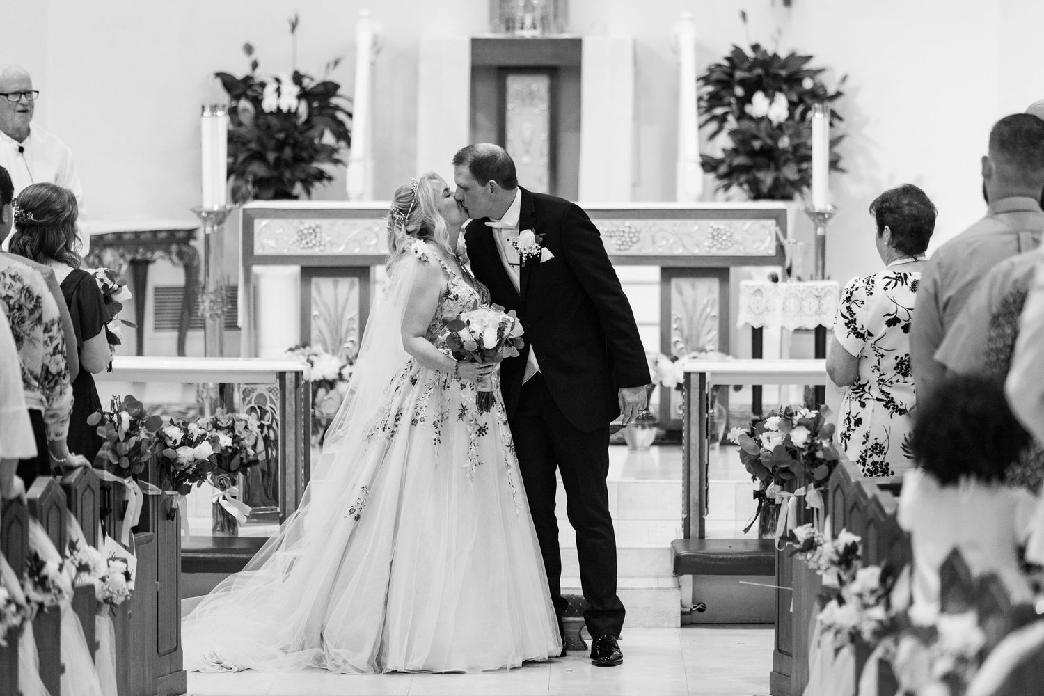 Black and white photo of newlyweds sharing first kiss at church altar after wedding ceremony.
