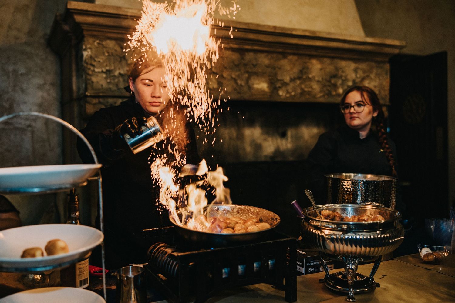 Catering staff performing flambé cooking display with dramatic flames at wedding reception dinner.