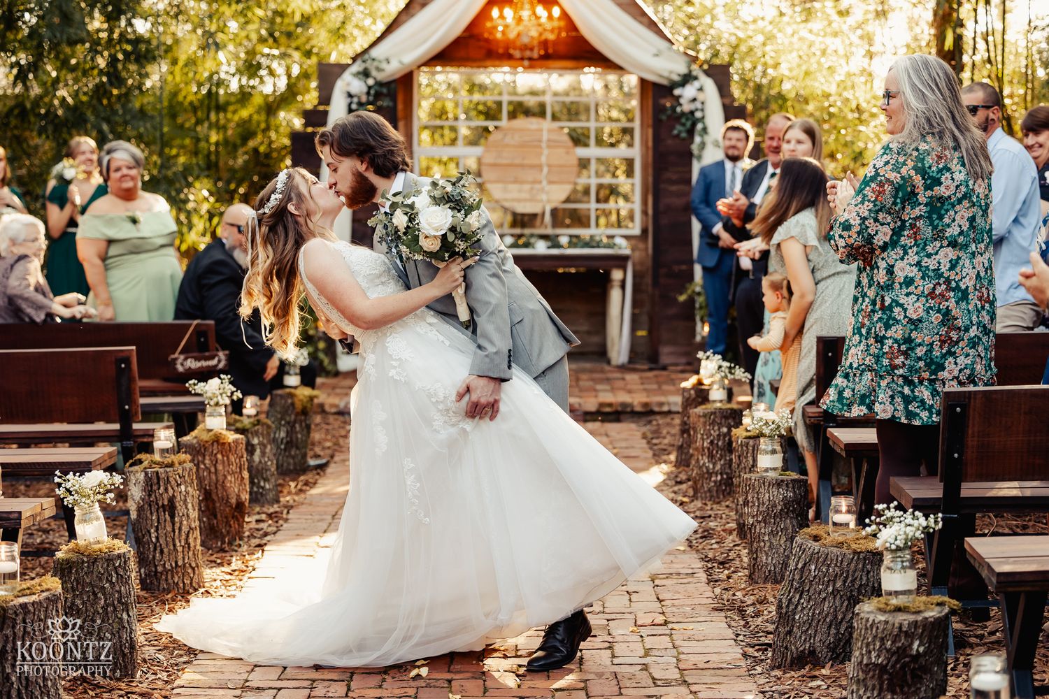 Bride and groom kiss on brick aisle lined with tree stumps and lanterns after outdoor ceremony.
