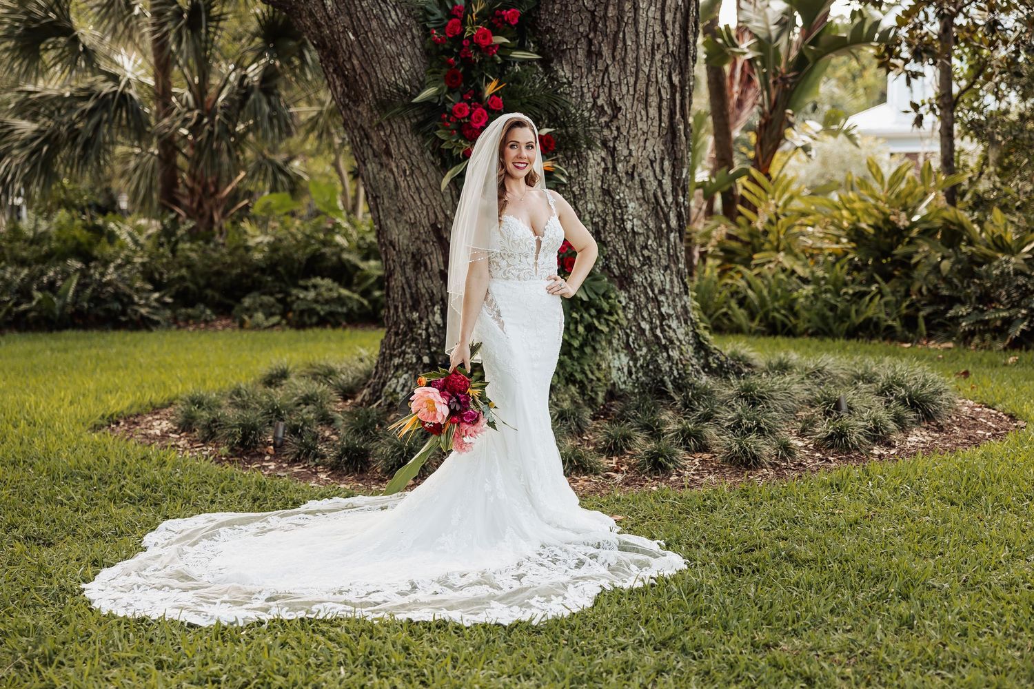 Bride in lace mermaid gown with long veil holds colorful bouquet beside large tree outdoors.