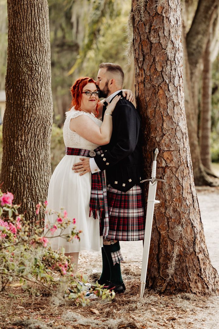Bride in white dress and groom in tartan kilt embracing against a pine tree with a sword nearby.