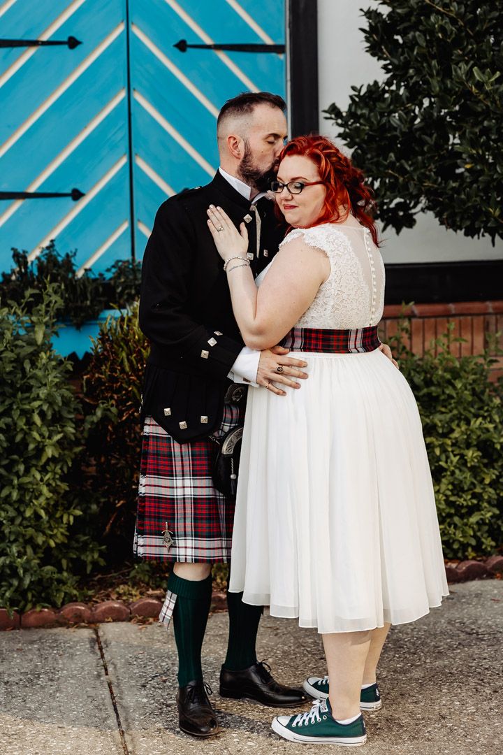 Groom in tartan kilt kissing red-haired bride wearing green Converse sneakers.
