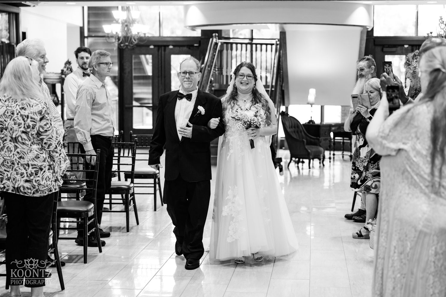 Wedding ceremony processional in an elegant indoor venue with guests looking on from wooden chairs.