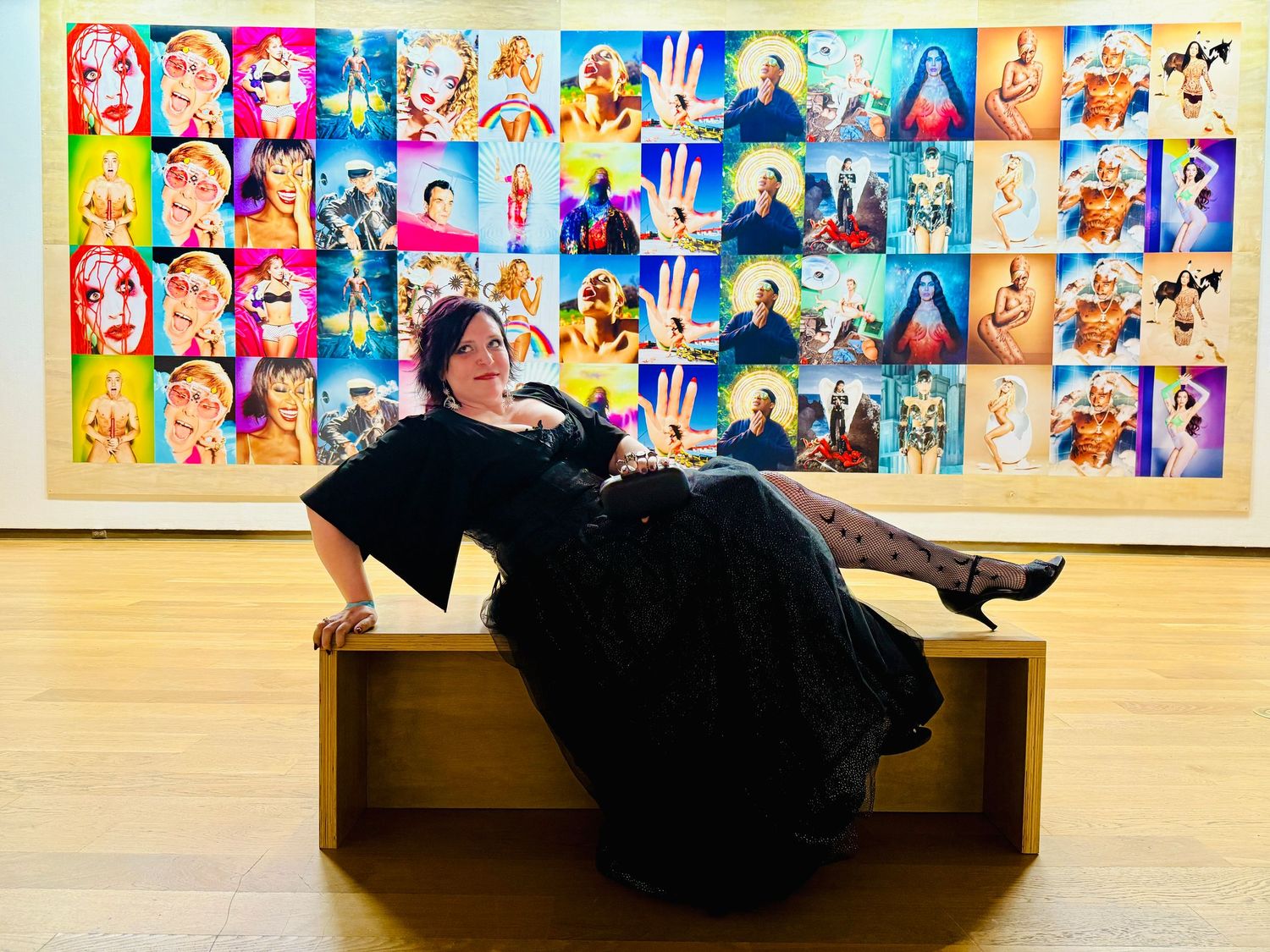 A person in black clothing lies down on a wooden bench against a colorful wall decorated with artwork.