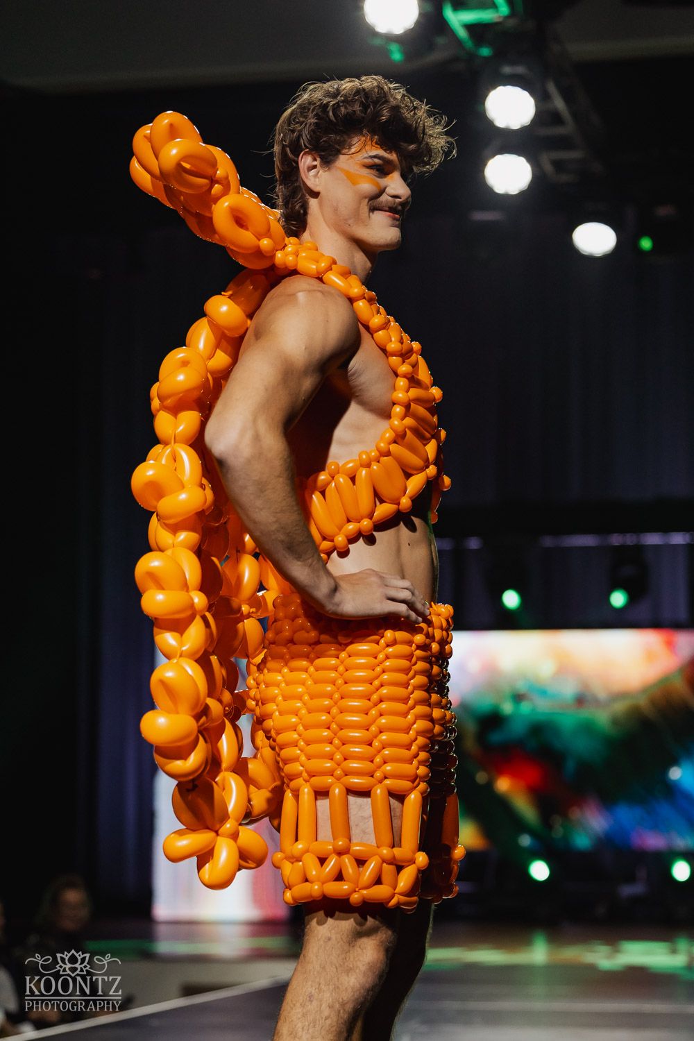 Model walks runway wearing avant-garde dress made entirely of bright orange balloons under dramatic stage lighting.
