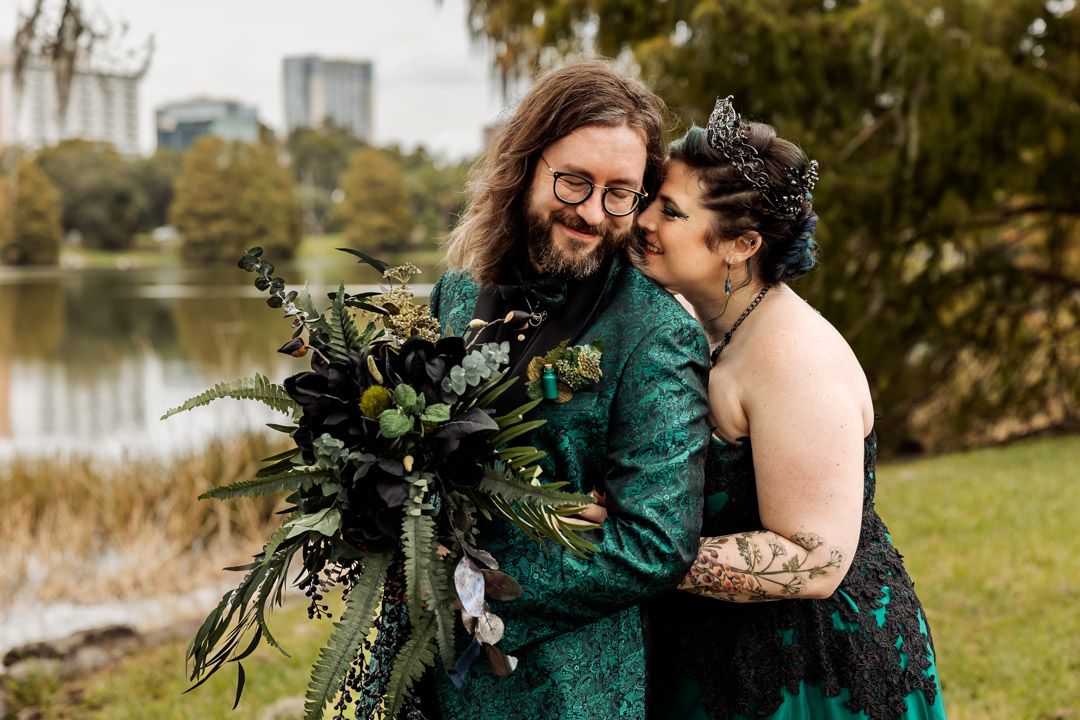 Two people in matching emerald green outfits embrace while holding a dark botanical bouquet by a lake on an autumn day.
