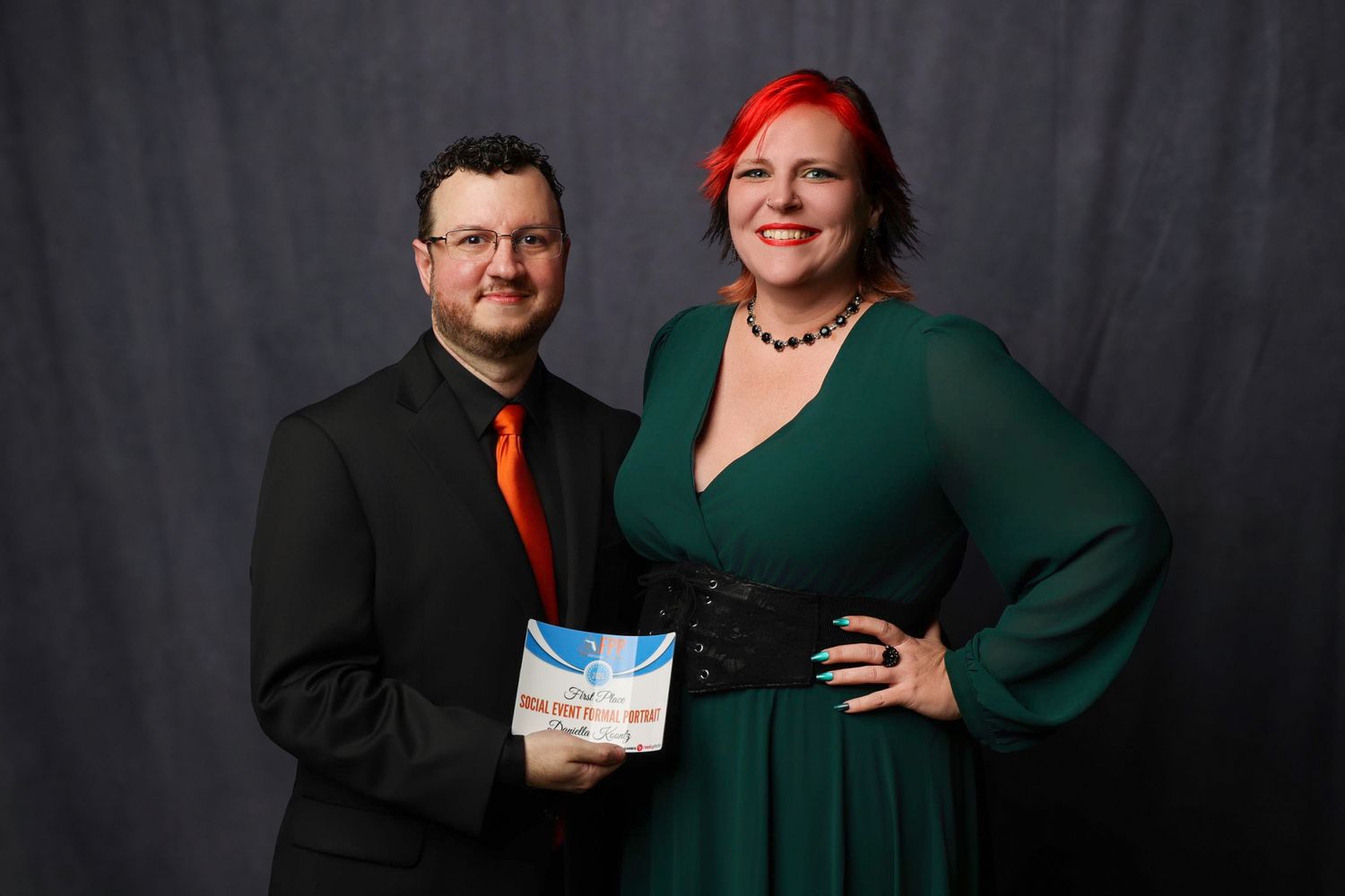 Couple in formal wear posing together at an awards ceremony, woman in green dress with red hair.