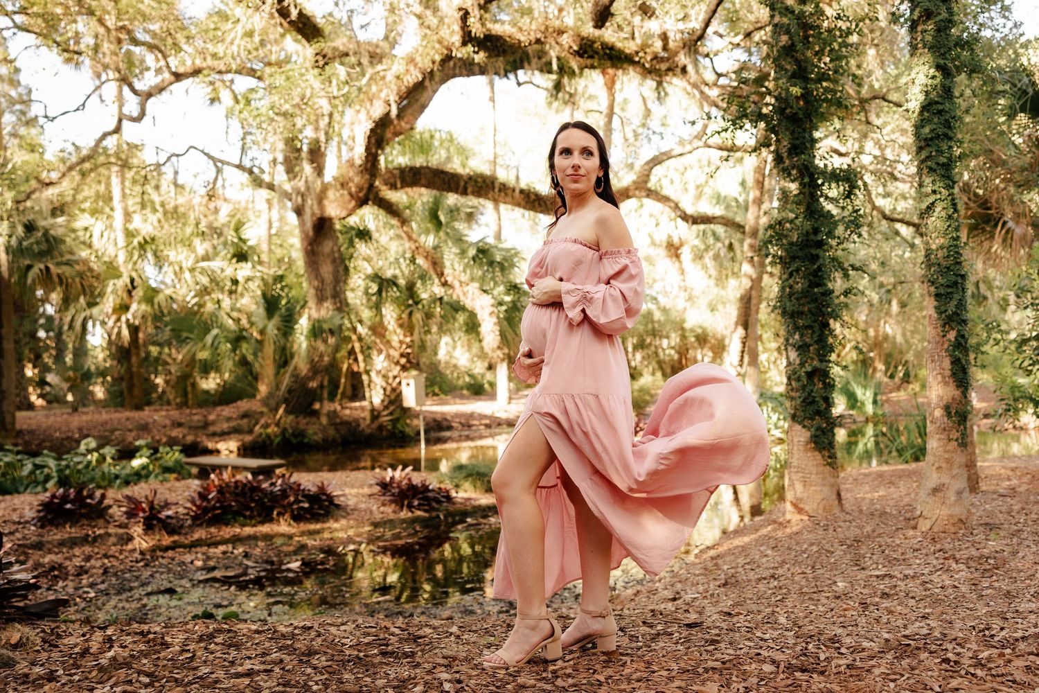 Pregnant woman in flowing pink off-shoulder dress posing gracefully among moss-draped oak trees in a lush garden.